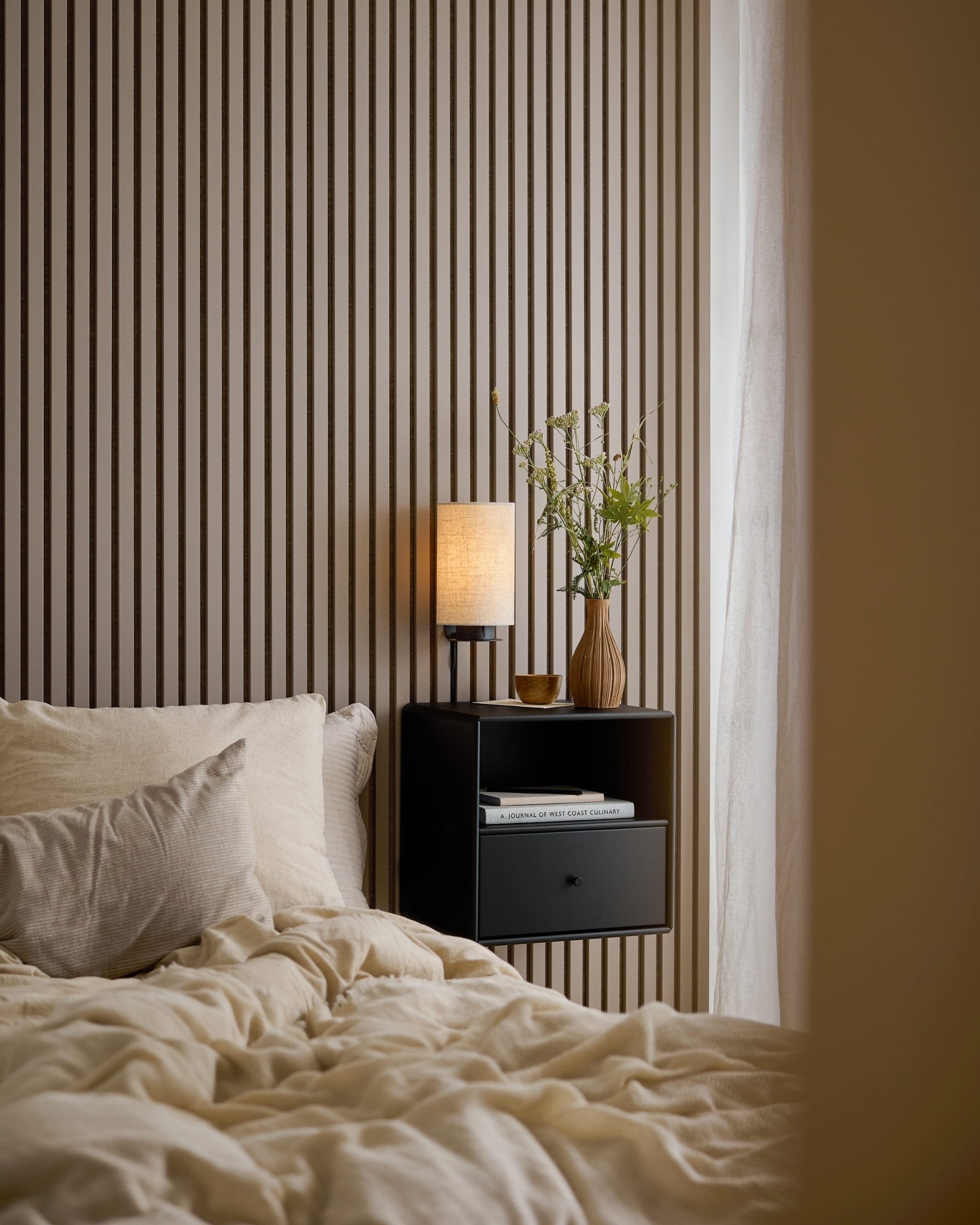 En säng med krämvita sängkläder bredvid ett svart nattduksbord, framför en Akupanel-vägg i Dark Cashmere.