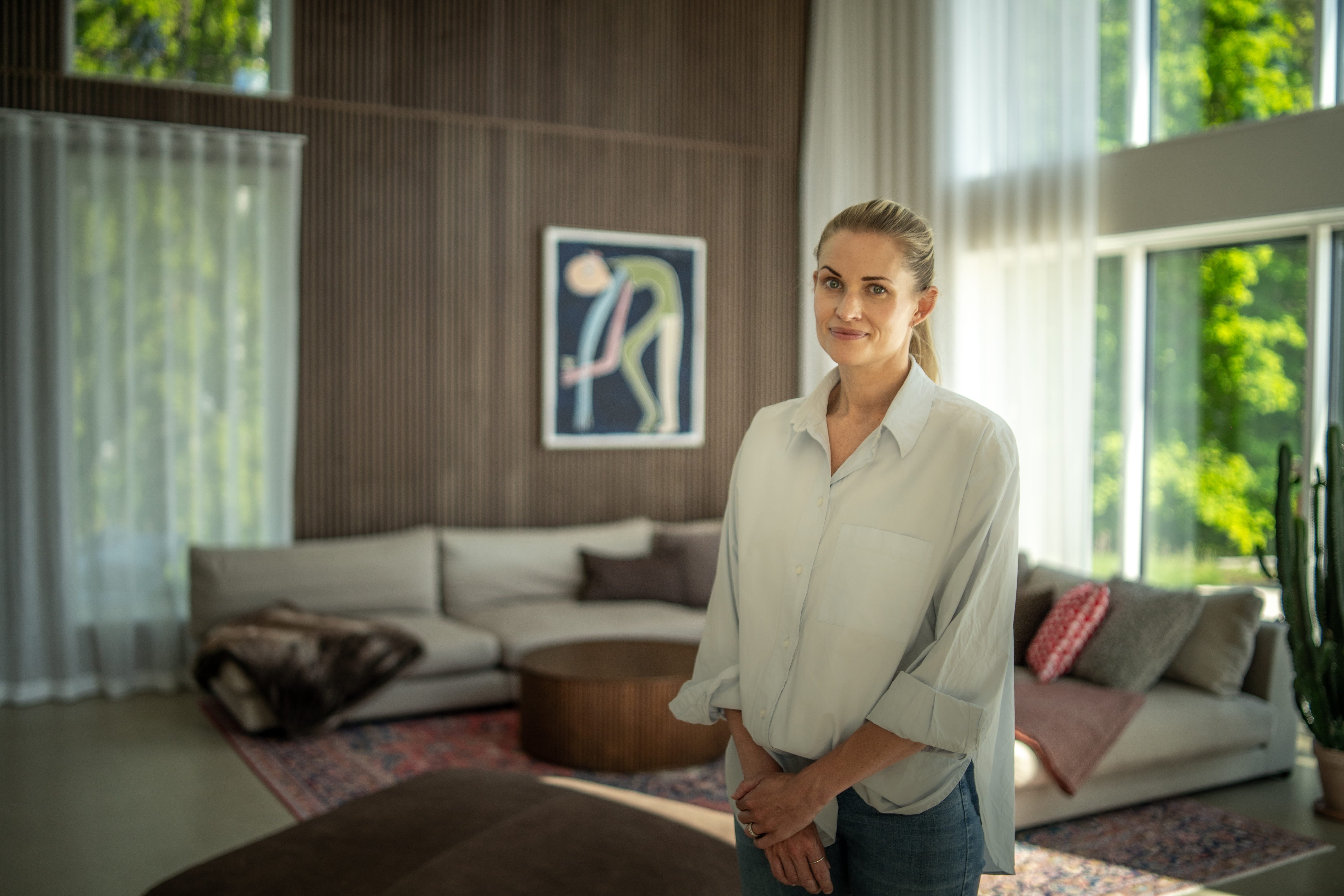 En kvinna står i ett solbelyst vardagsrum med stora fönster, nordiska akustiska träpaneler på väggen, modern konst och minimalistiska möbler i neutrala toner.
