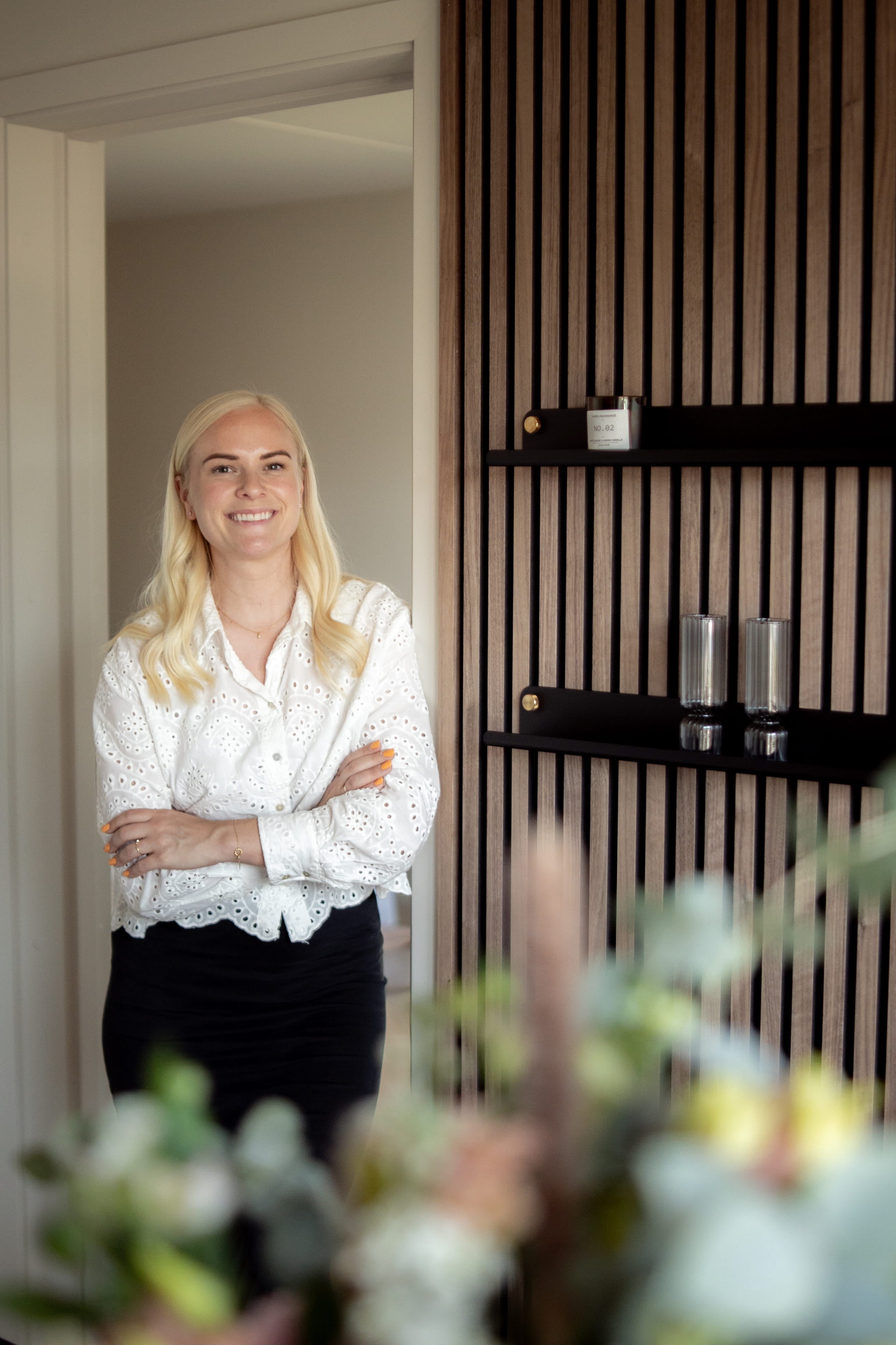 En kvinna med långt blont hår och vit blus står leende med armarna i kors i ett modernt rum med nordisk akustisk träpanel och snygga hyllor.