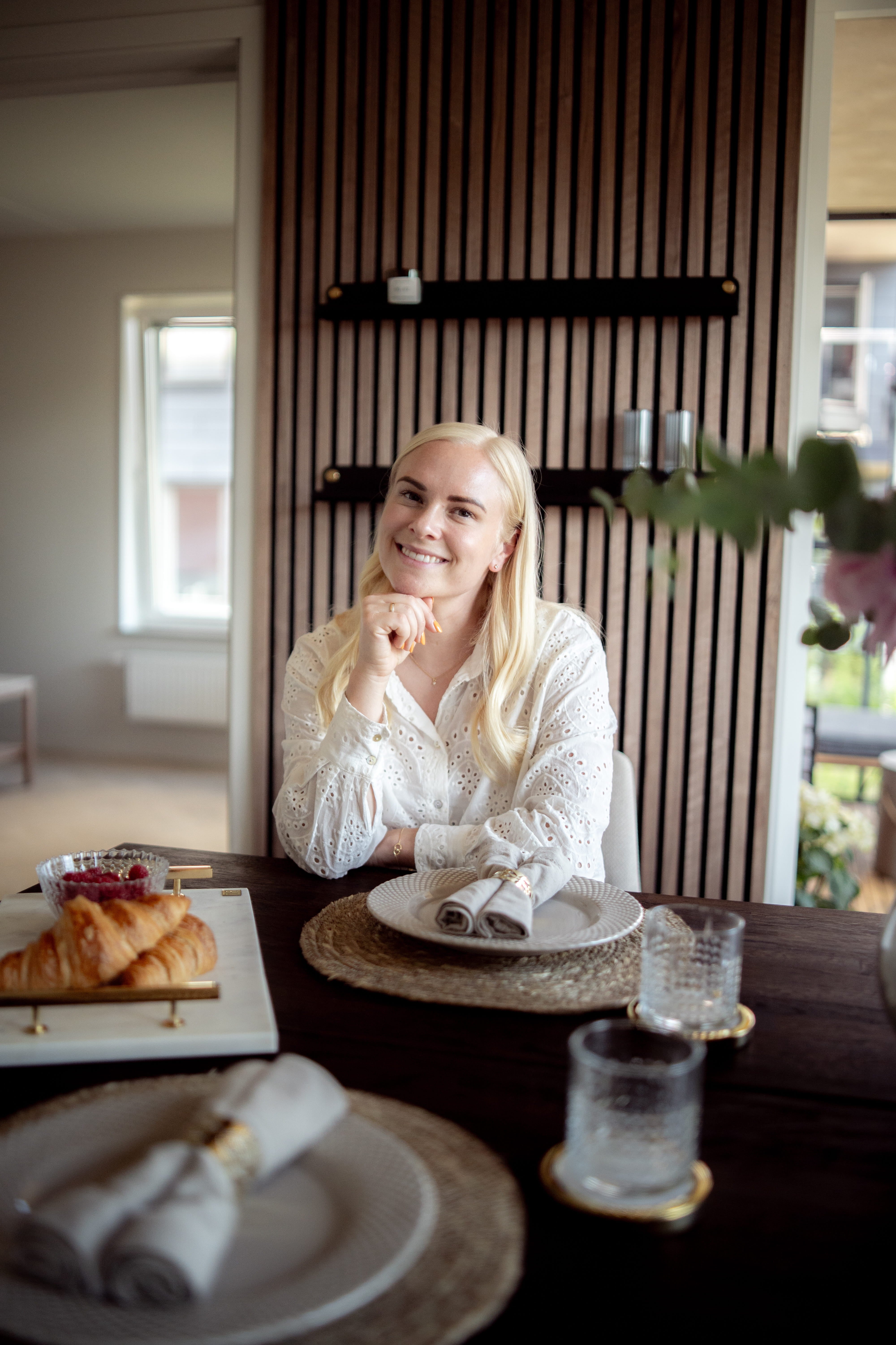 En kvinna med långt blont hår sitter vid ett matbord dukat för frukost med croissanter och glas, bredvid en nordisk akustikpanel på väggen.