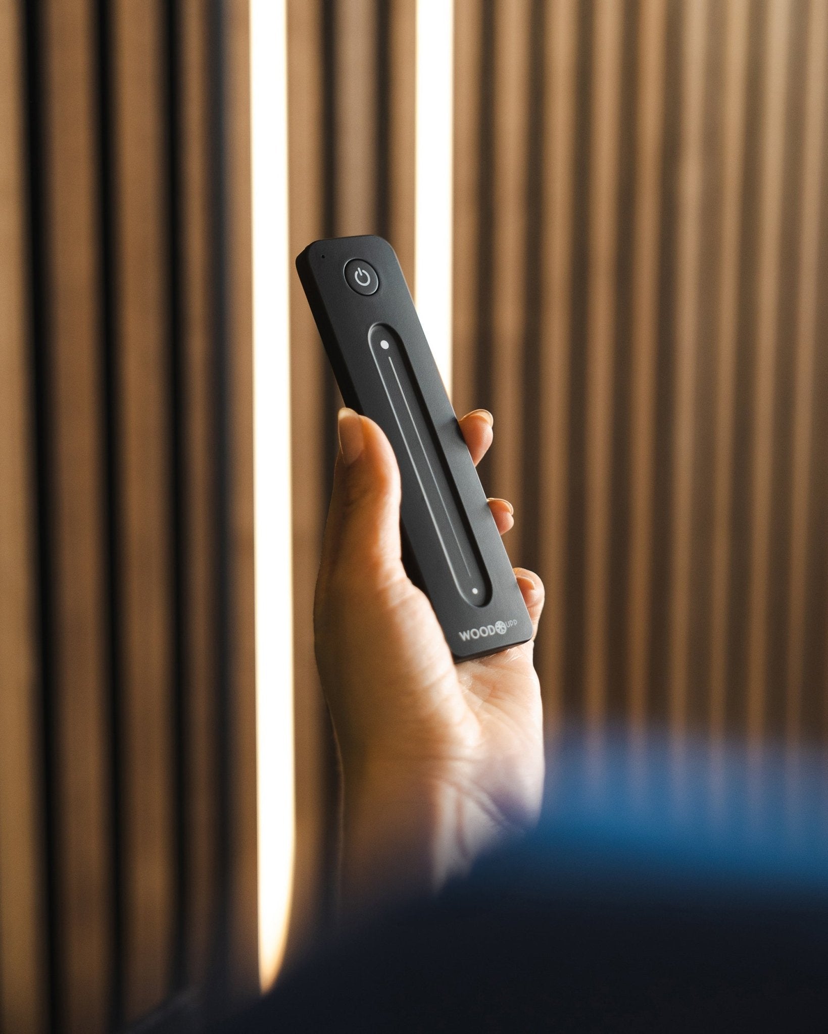 En hand håller en black WoodUpp Lumi-fjärrkontroll för LED-lister med strömknapp, placerad framför WoodUpps akustiska ribbpaneler; kompatibel med Philips Hue.