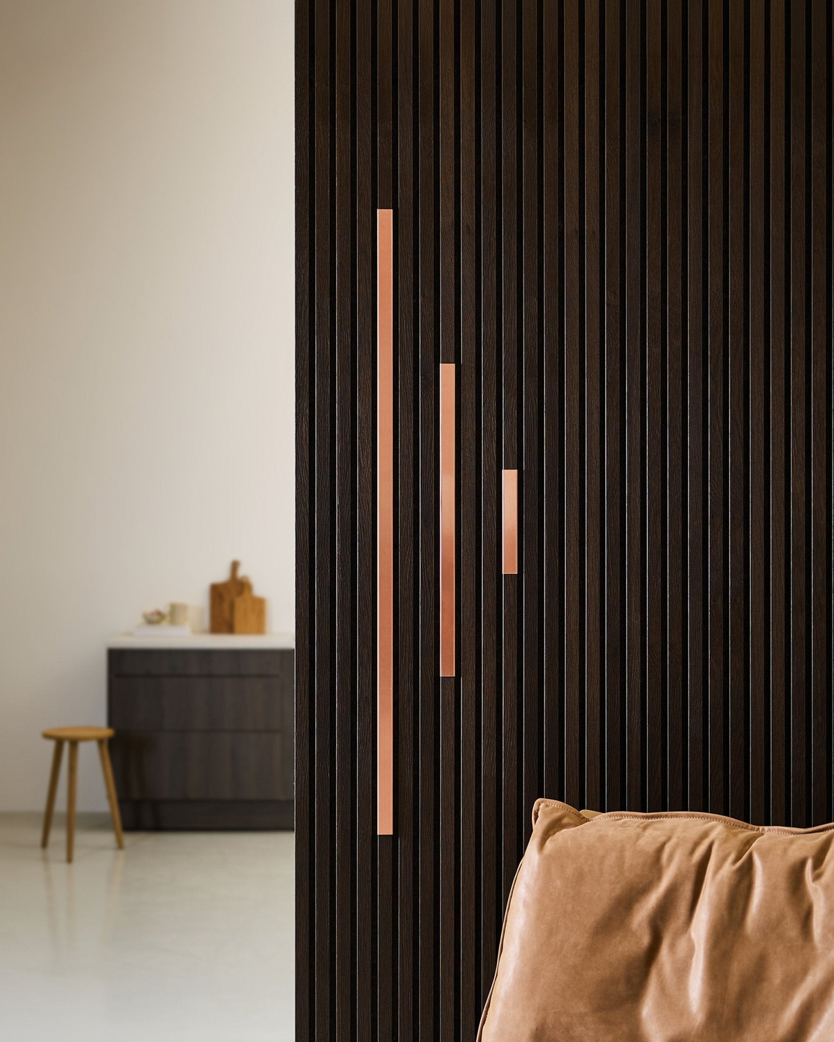 Minimalistisk miljö med en mörk akustisk ribbvägg i trä med WoodUpp Liva Cover i copper, 60 cm paneler med copper handtag, en stol i ljusbrunt läder och en låg skåpspall.