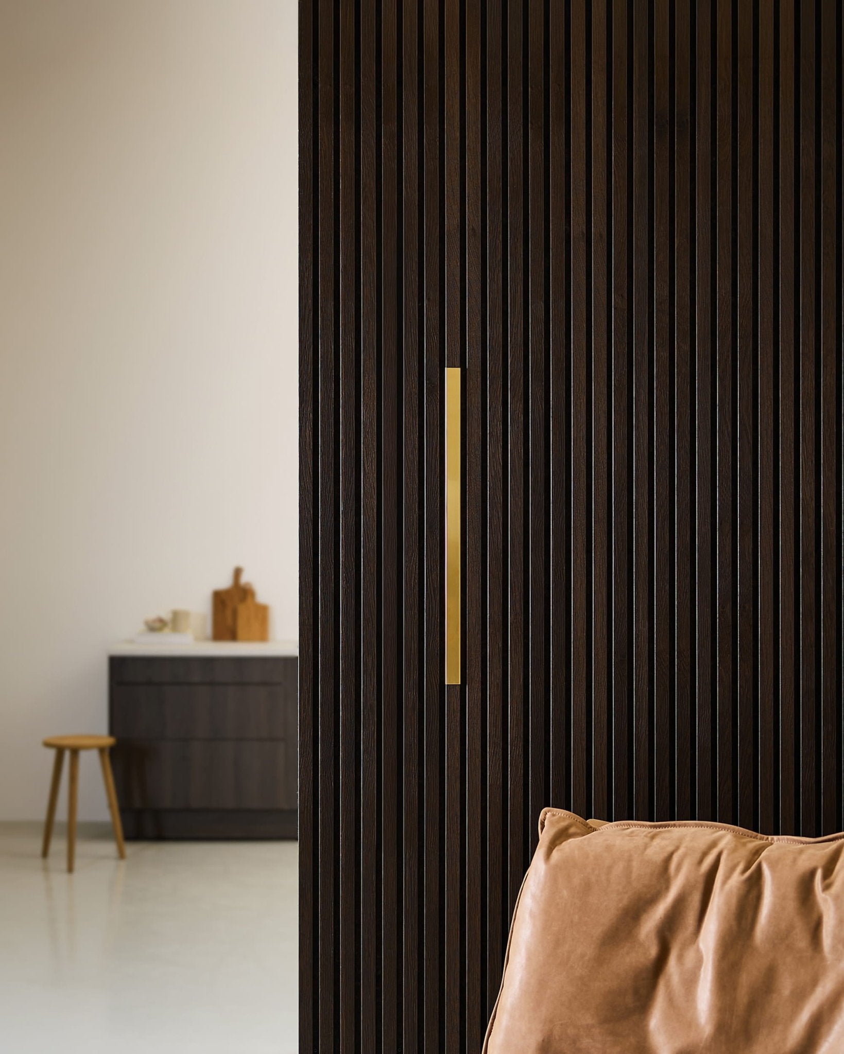Ett minimalistiskt rum med en WoodUpp Akupanel Liva | Cover / mässing, 60 cm, bredvid en läderfåtölj, en pall och ett mörkt skåp i en neutral miljö.