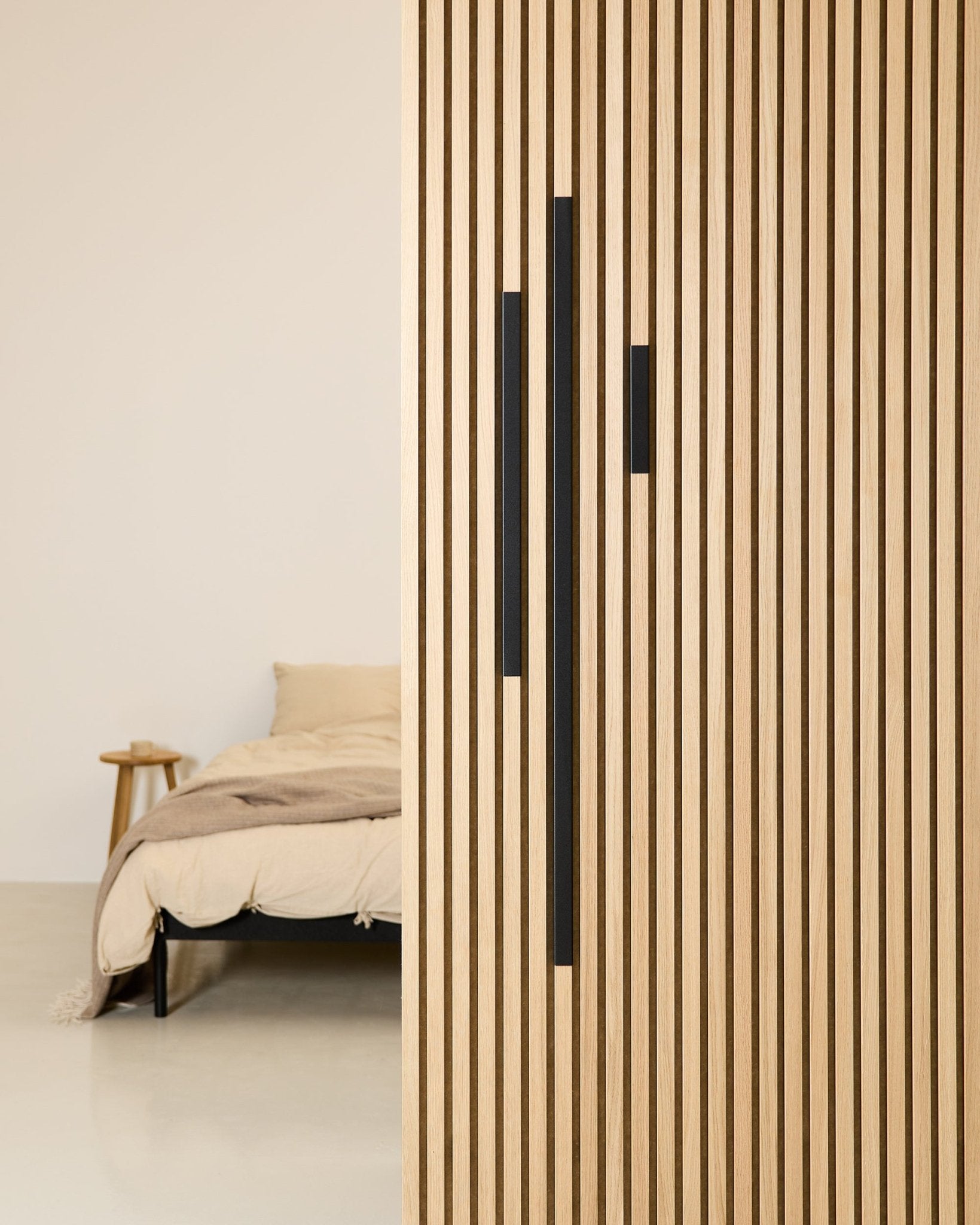 Minimalistiskt sovrum med en beige säng och filt, delvis skymt av en 60 cm WoodUpp Akupanel rumsavdelare i Black aluminium från serien Liva Cover.