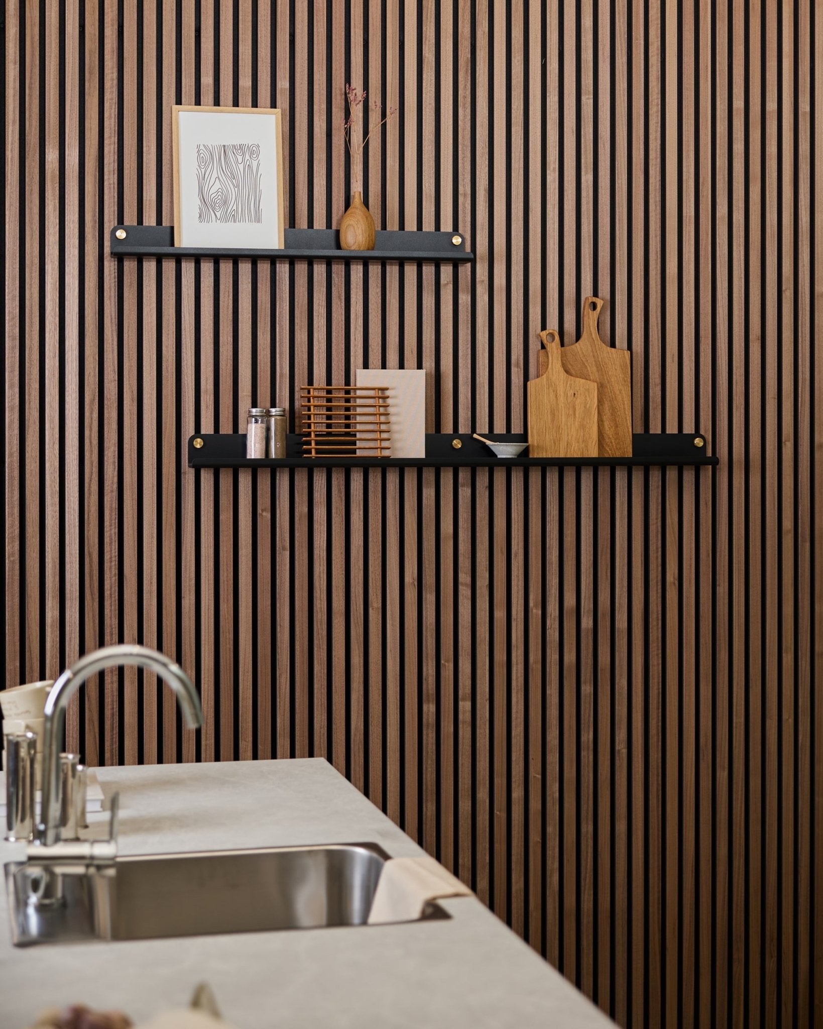 Minimalistiskt kök med en vertical Akupanel akustisk ribbvägg i trä, black hyllor, skärbrädor, small inredningsdetaljer och en modern diskbänk framför.