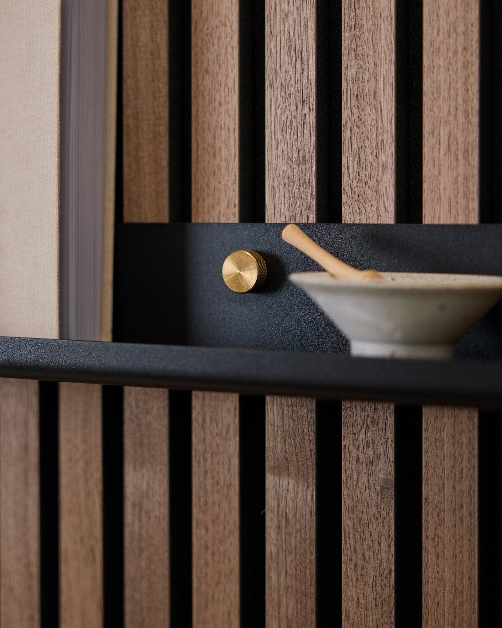 Närbild av en 115,9 cm black WoodUpp Norr Narrow Shelf monterad på en nordisk akustisk ribbväggspanel med brass tumskruvar, gyllene knopp och keramisk skål med sked.