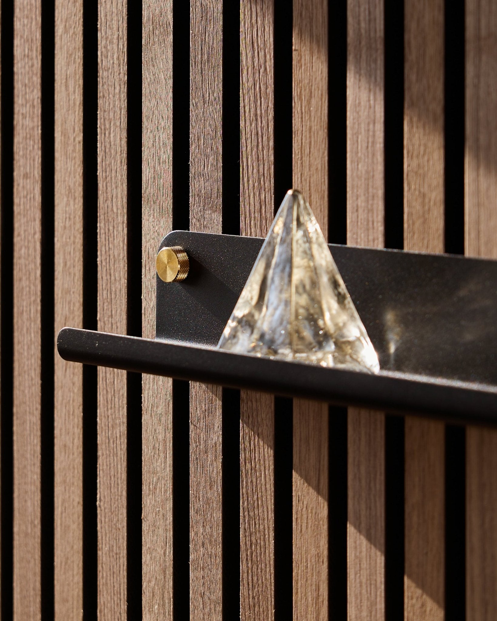 En glaspyramid står på WoodUpp Norr Narrow Shelf i black (51,9 cm), monterad på en vertical Akustikpanel och framhävd av solljus.