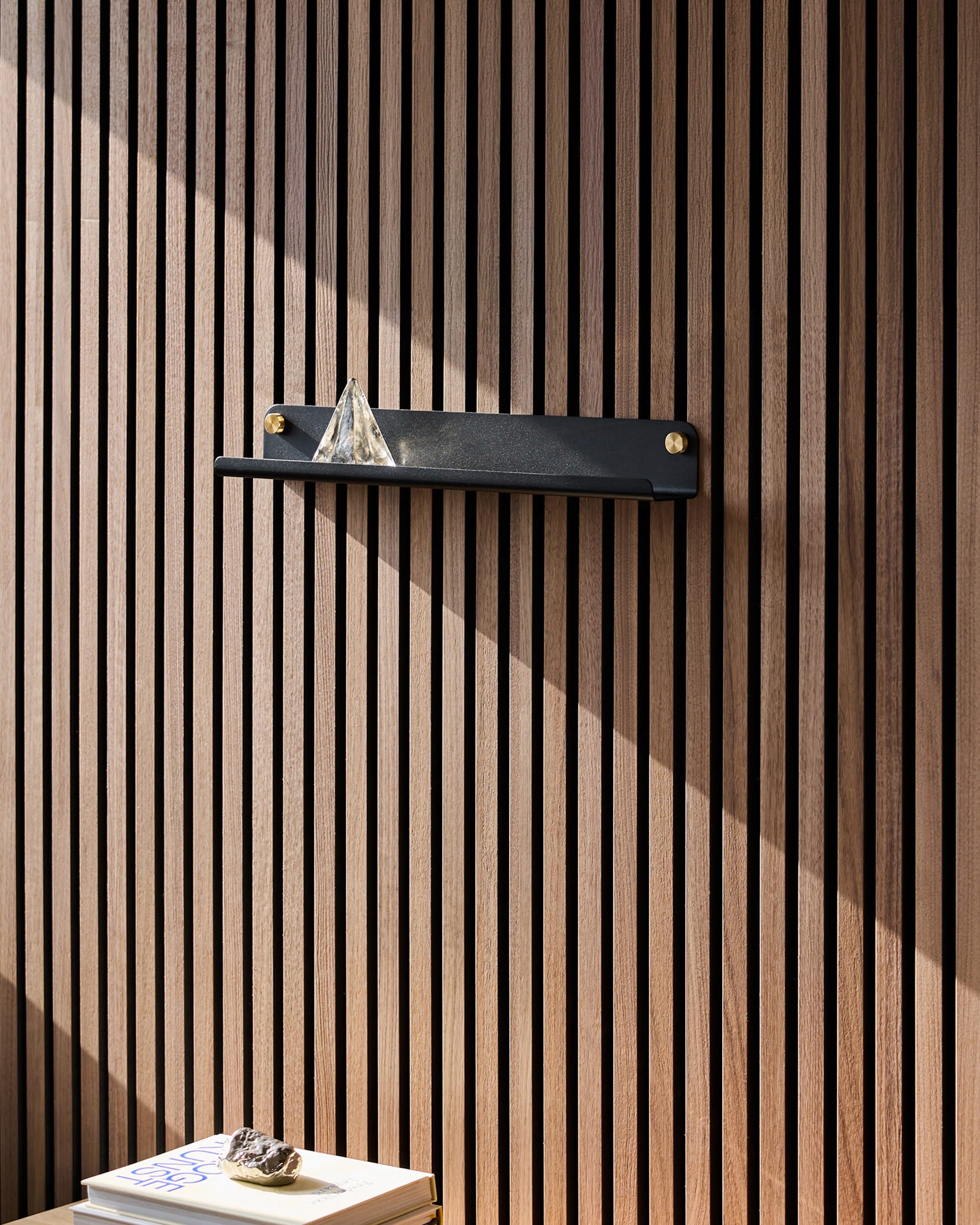 En svart Norr Narrow Shelf från WoodUpp, placerad på en akustisk träpanel från Akupanel, bär upp en glaspyramid; solljuset faller på böckerna nedanför, där en sten ligger ovanpå.
