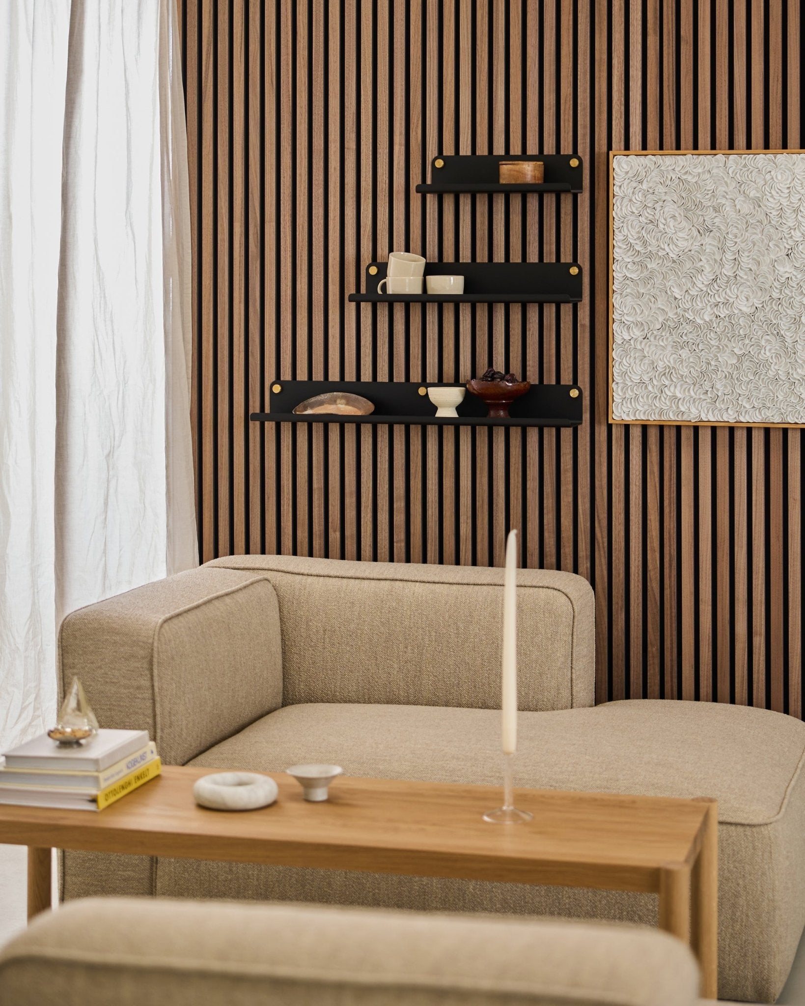 Modern living room with a beige sofa, wooden table, black WoodUpp Norr Wide Shelf (51.9 cm), and a textured Akupanel acoustic panel wall featuring abstract art.
