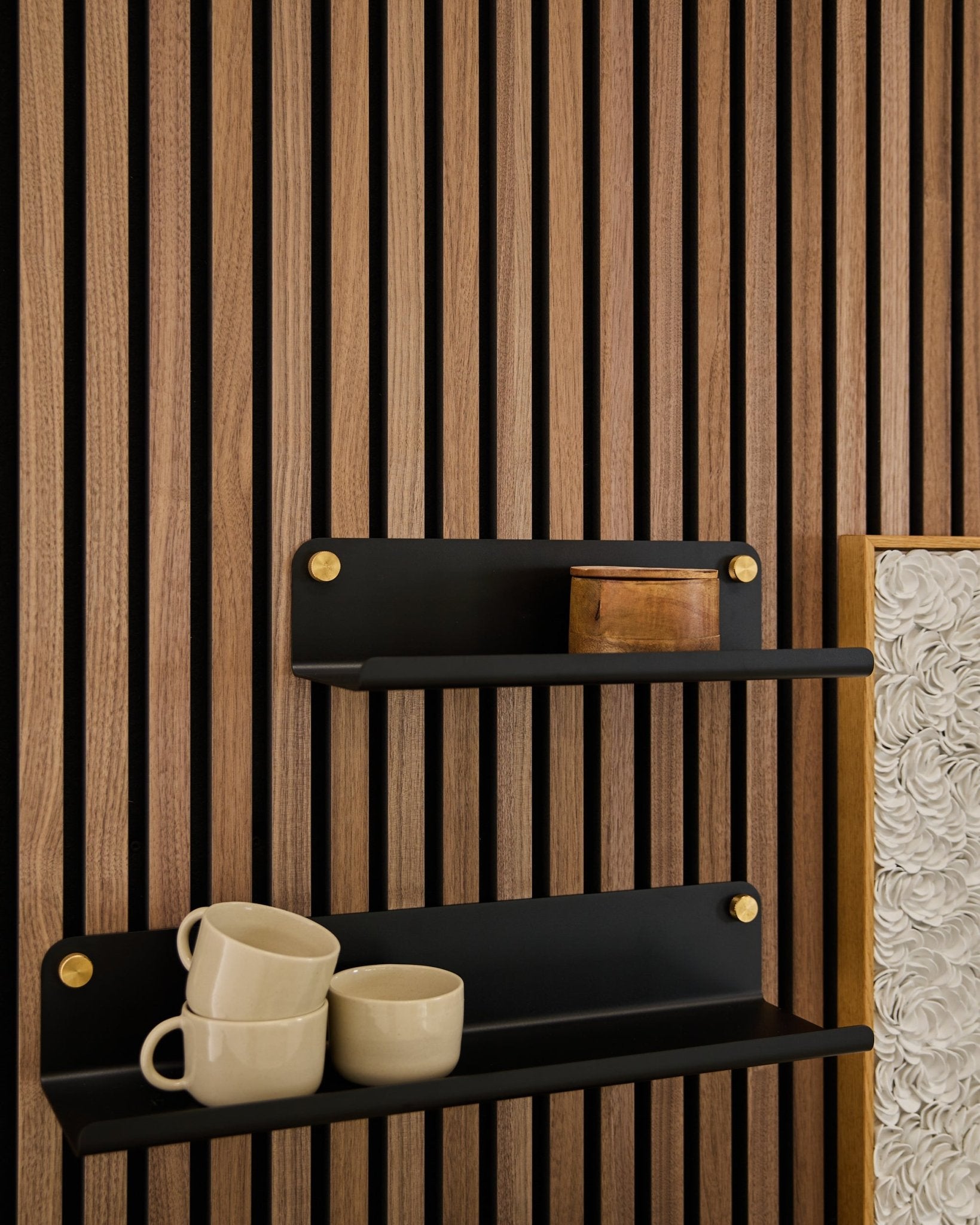 Två black Norr | Wide Shelves från WoodUpp (35,9 cm) med guldfärgade konsoler monterade på en Akupanel akustikpanel i trä; den nedre hyllan har 3 beige muggar, den övre har en small trälåda.