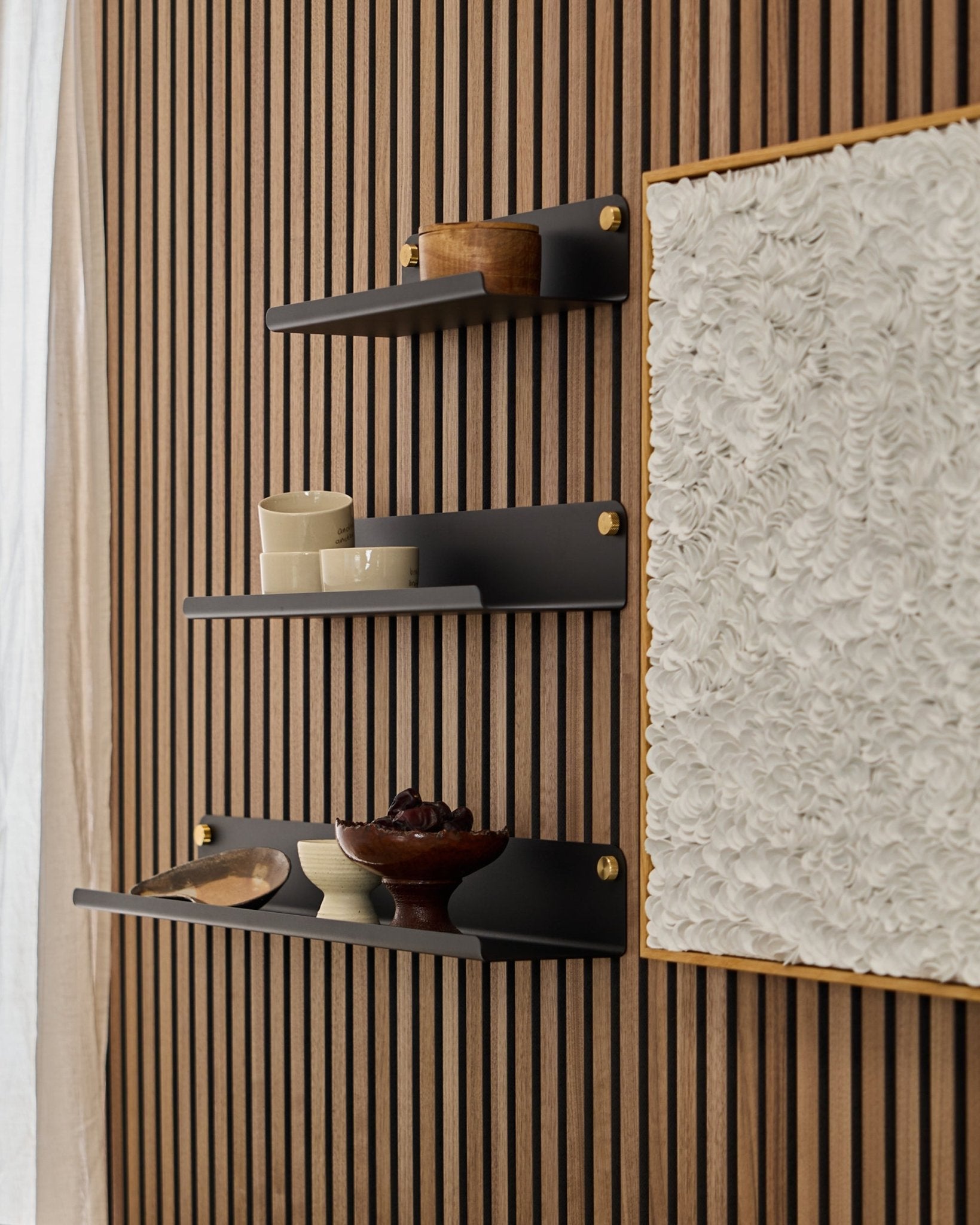 Tre black WoodUpp Norr Wide Shelves (51,9 cm) monterade på en WoodUpp Akupanel akustikträpanel, bredvid texturerad vit konst, med skålar och träbehållare på.