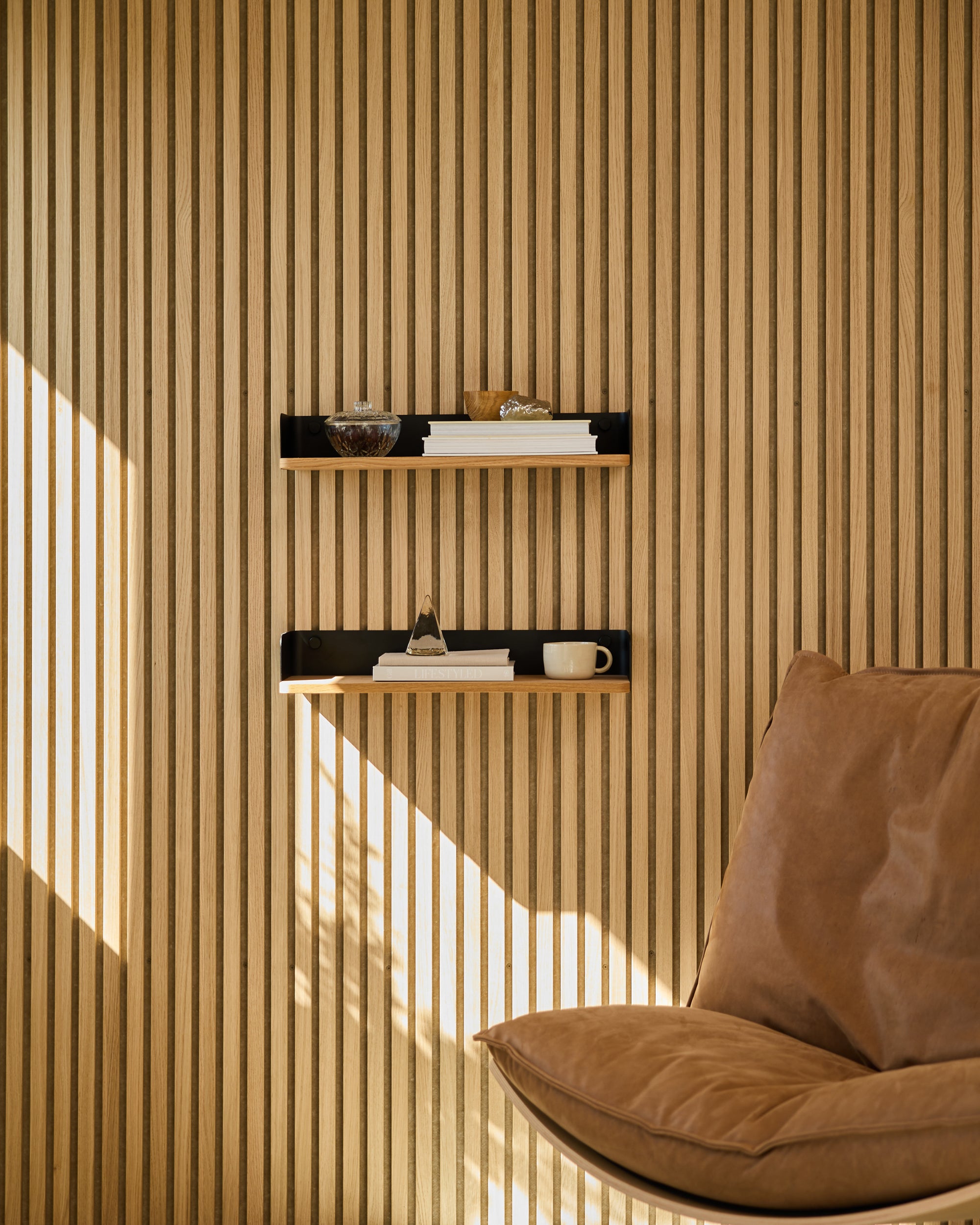 Modern Nordic acoustic wood panel wall with minimalist shelves holding books and decor, next to a tan leather chair illuminated by sunlight.