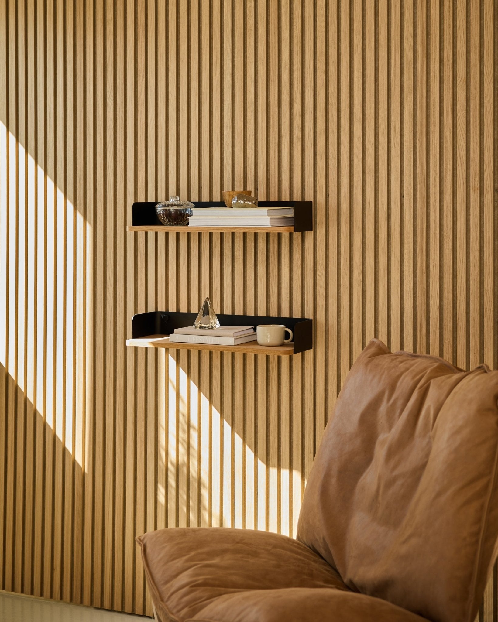 Brown stol bredvid WoodUpp Linn bokhylla i Natural Oak, med böcker och inredningsdetaljer arrangerade i naturligt ljus intill en nordisk vägg med trälameller Akustikpanel.