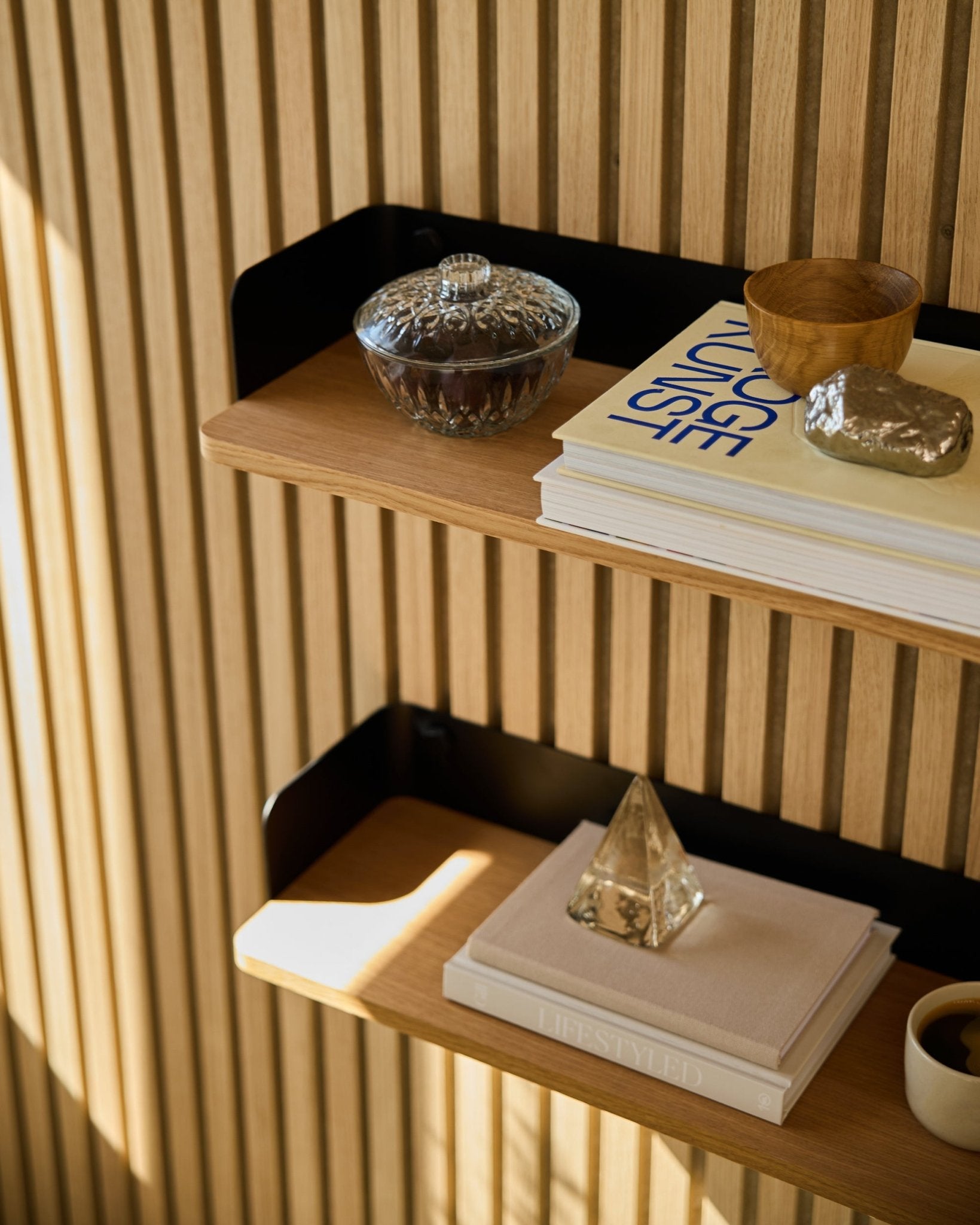 WoodUpp Linn Bookshelf i Natural Oak (55,9 cm) rymmer en glasburk, kopp, guldfärgad pyramid och böcker framför Akupanel akustikpaneler i trä i solljus.