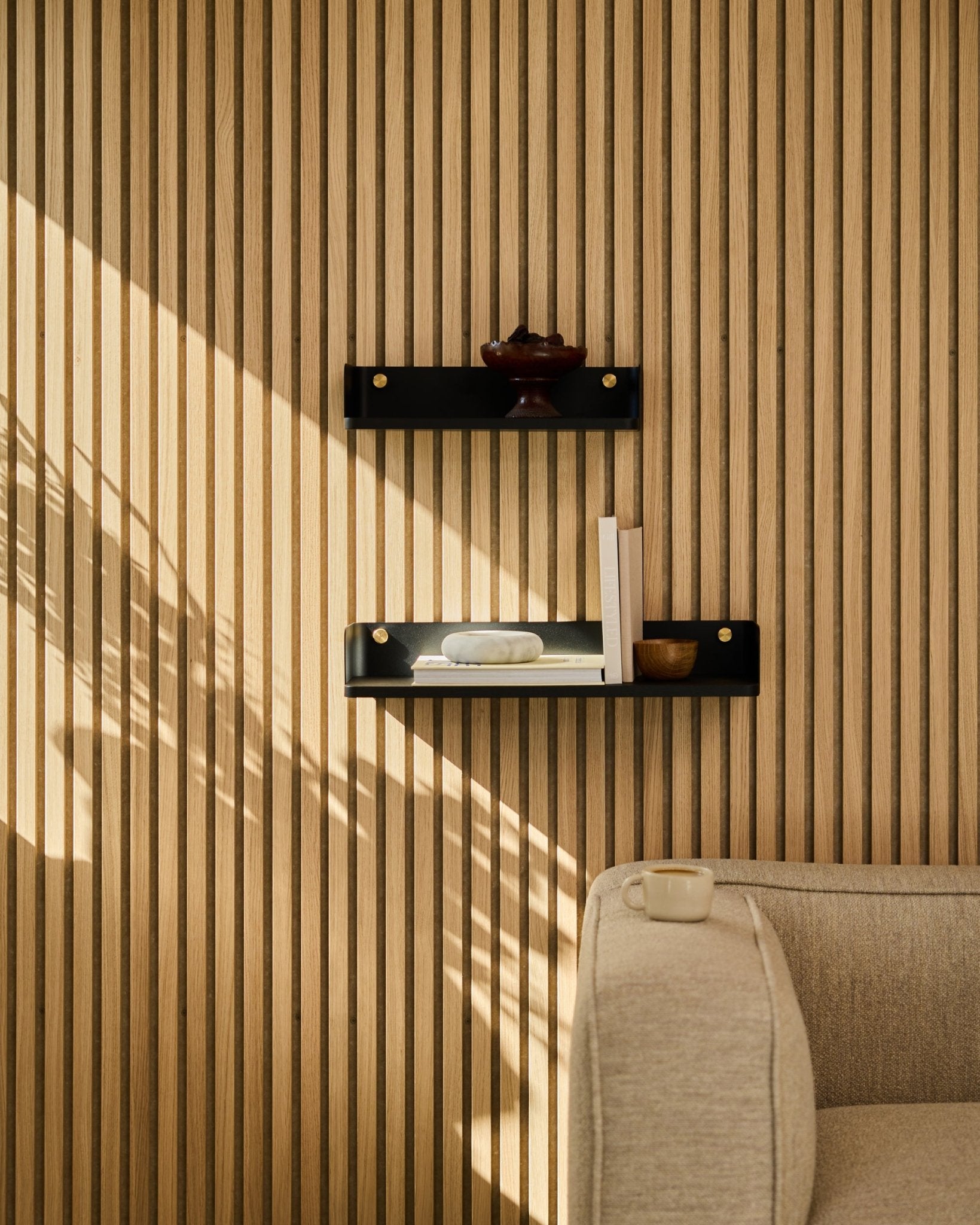 Solljuset lyser upp en soffa i beige och WoodUpps bokhylla Linn i black oak, 55,9 cm, mot en minimalistisk nordisk ribbvägg Akustikpanel.