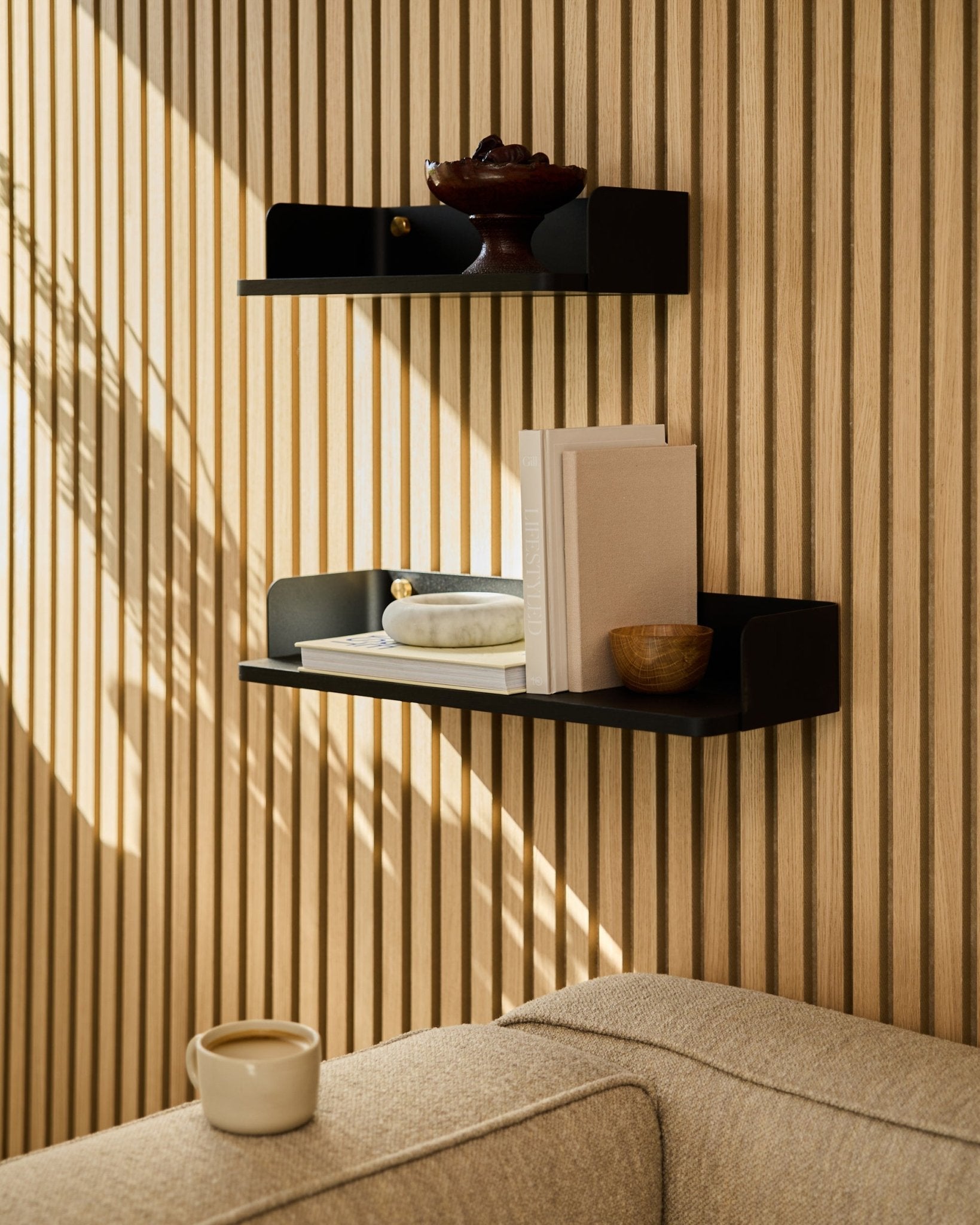 En WoodUpp-bokhylla i black oak (55,9 cm) visar böcker, dekoration och en skål på en vägg med akustisk träpanel; en soffa i beige och en mugg syns i förgrunden.