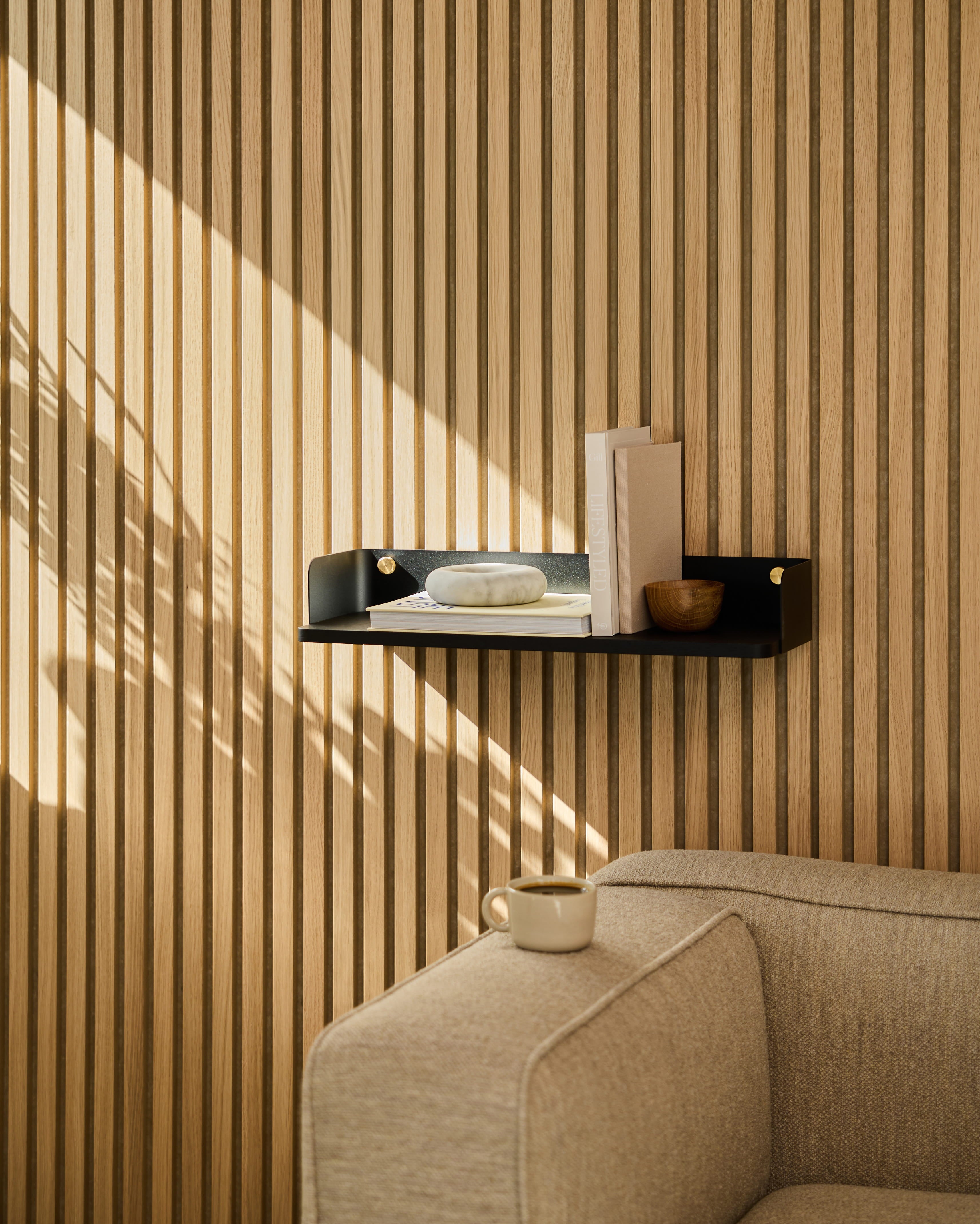En Linn | Bokhylla / Black Oak från WoodUpp pryder väggen ovanför en beige soffa. Solljuset kastar skuggor på den randiga akustikpanelen av trä.