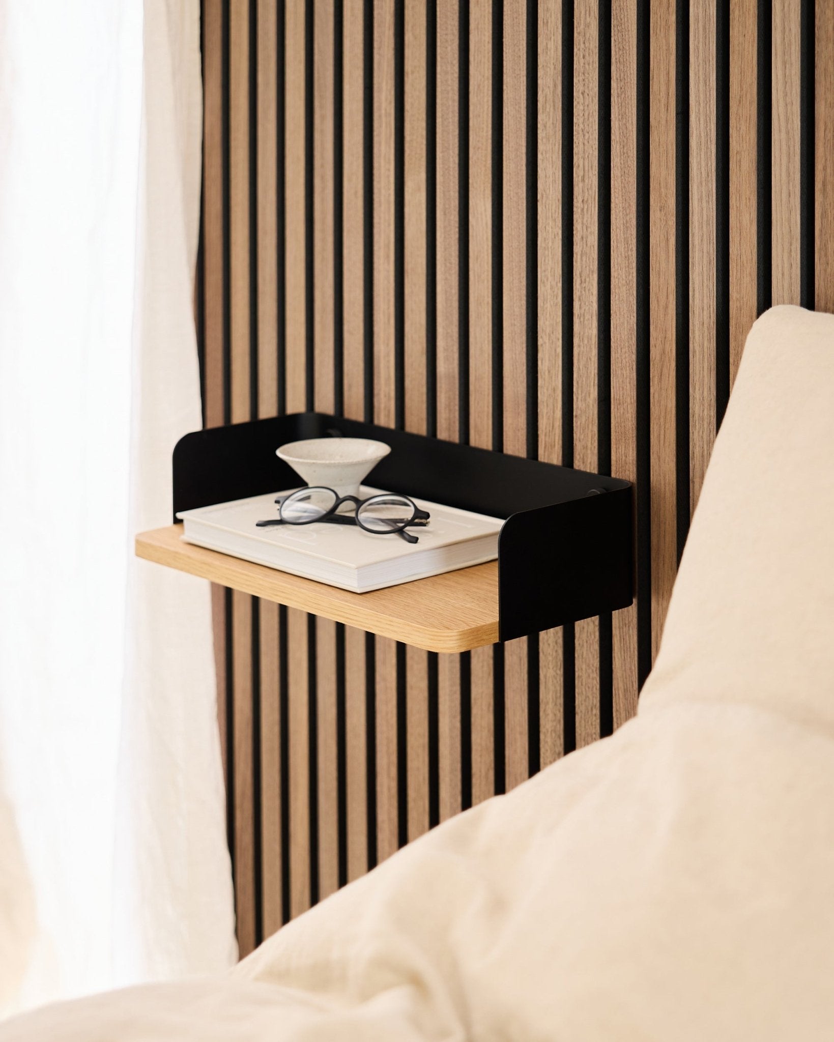 Linn bokhylla från WoodUpp i natural oak (39,9 cm) med en bok, glasögon och ett glas bredvid en säng mot en vägg med Akupanel akustikpanel i trä.