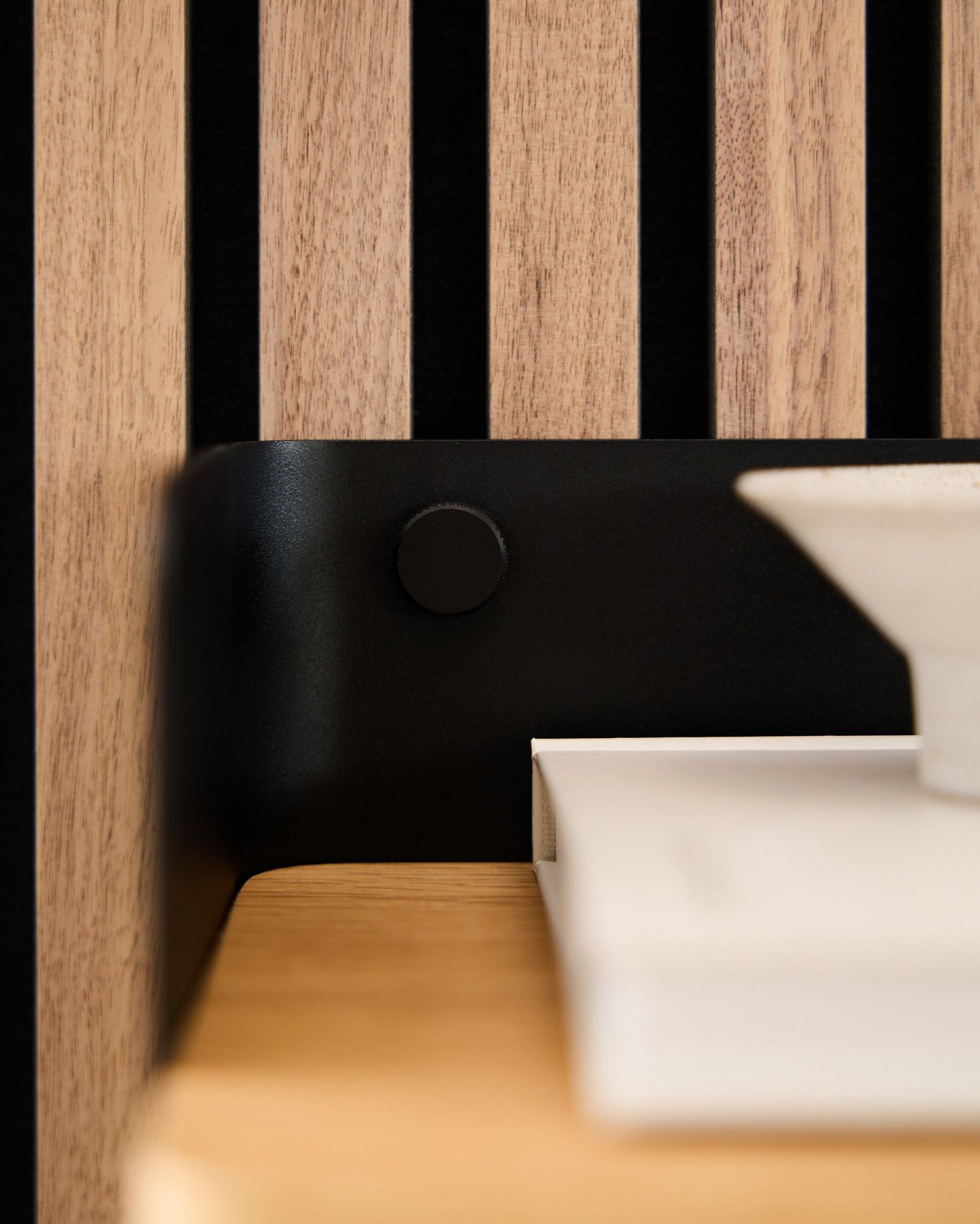 Närbild av en WoodUpp Akupanel akustikpanel med träribbor där black Finger-skruvar fäster ett black fäste, tillsammans med vita böcker och en skål.