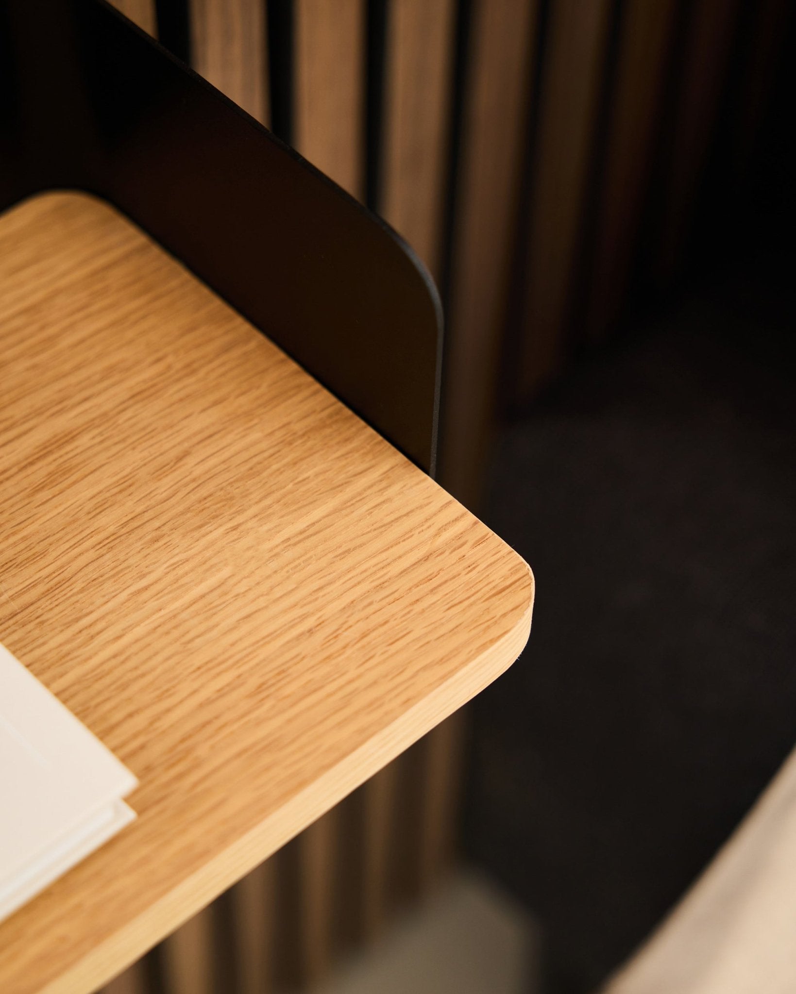 Närbild av ett skrivbordshörn med black Akupanel akustikpanel i trä, vita papper och WoodUpp Linn bokhylla i Natural Oak (39,9 cm) i bakgrunden.