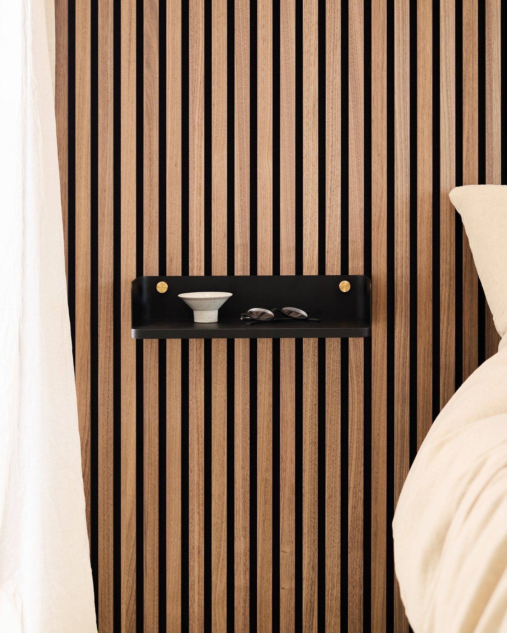 Linn Bokhylla i Black Oak från WoodUpp är monterad på en nordisk akustikpanel med ribbor och rymmer en skål och glas bredvid en säng med beige sängkläder.