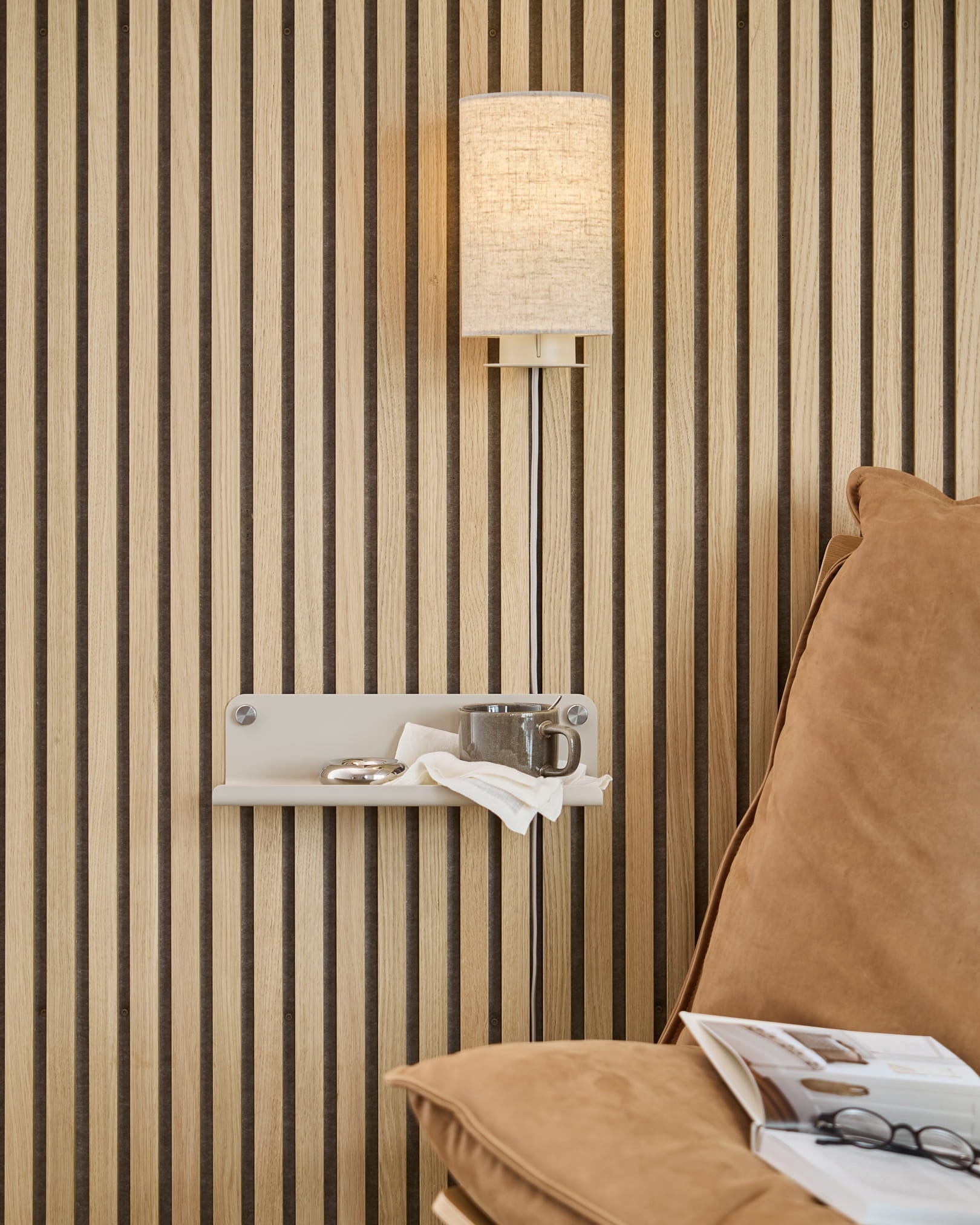 Vertikal Akupanel-vägg med akustiska trälister, beige vägglampa, liten hylla med kopp och servett, beige soffa med en öppen tidning.