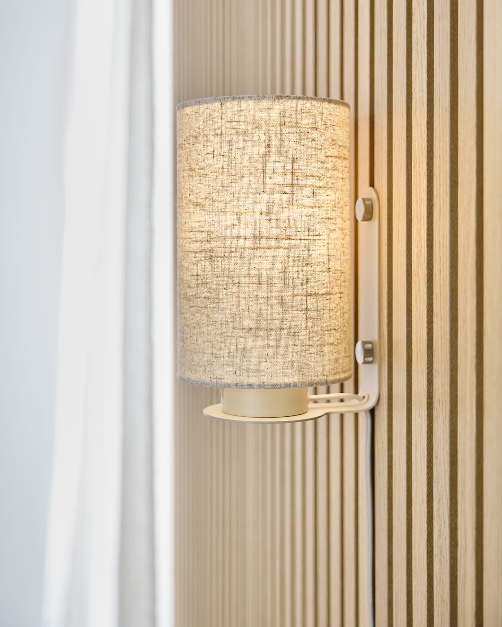 Feya vägglampa i Beige från WoodUpp sprider ett varmt ljus över en beige Akupanel akustisk ribbvägg, placerad intill en vit gardin och en skärm i linne-bomull.