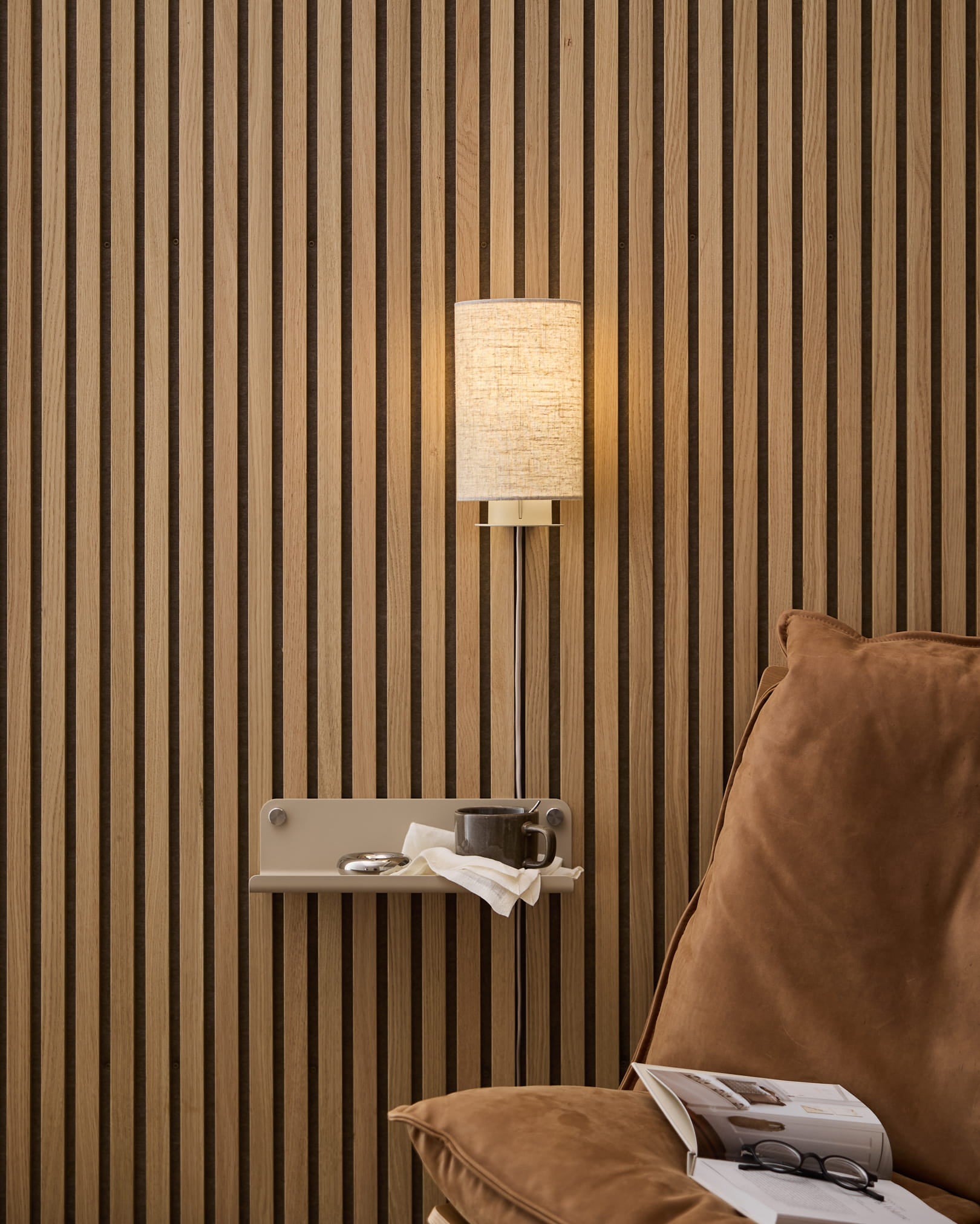 En beige Feya vägglampa i linne-bomull från WoodUpp monterad ovanför en inbjudande fåtölj och ett sidobord med bok och kopp, mot en nordisk ribbvägg i trä Akustikpanel.
