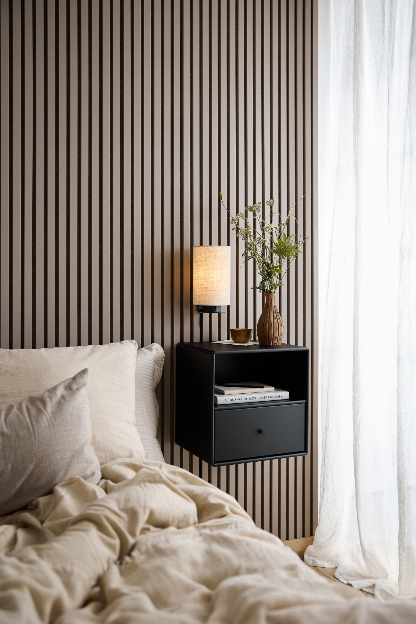 Säng med beige sängkläder, svart WoodUpp Arka bokhylla med låda, lampa och vas intill ett fönster med tunna vita gardiner.
