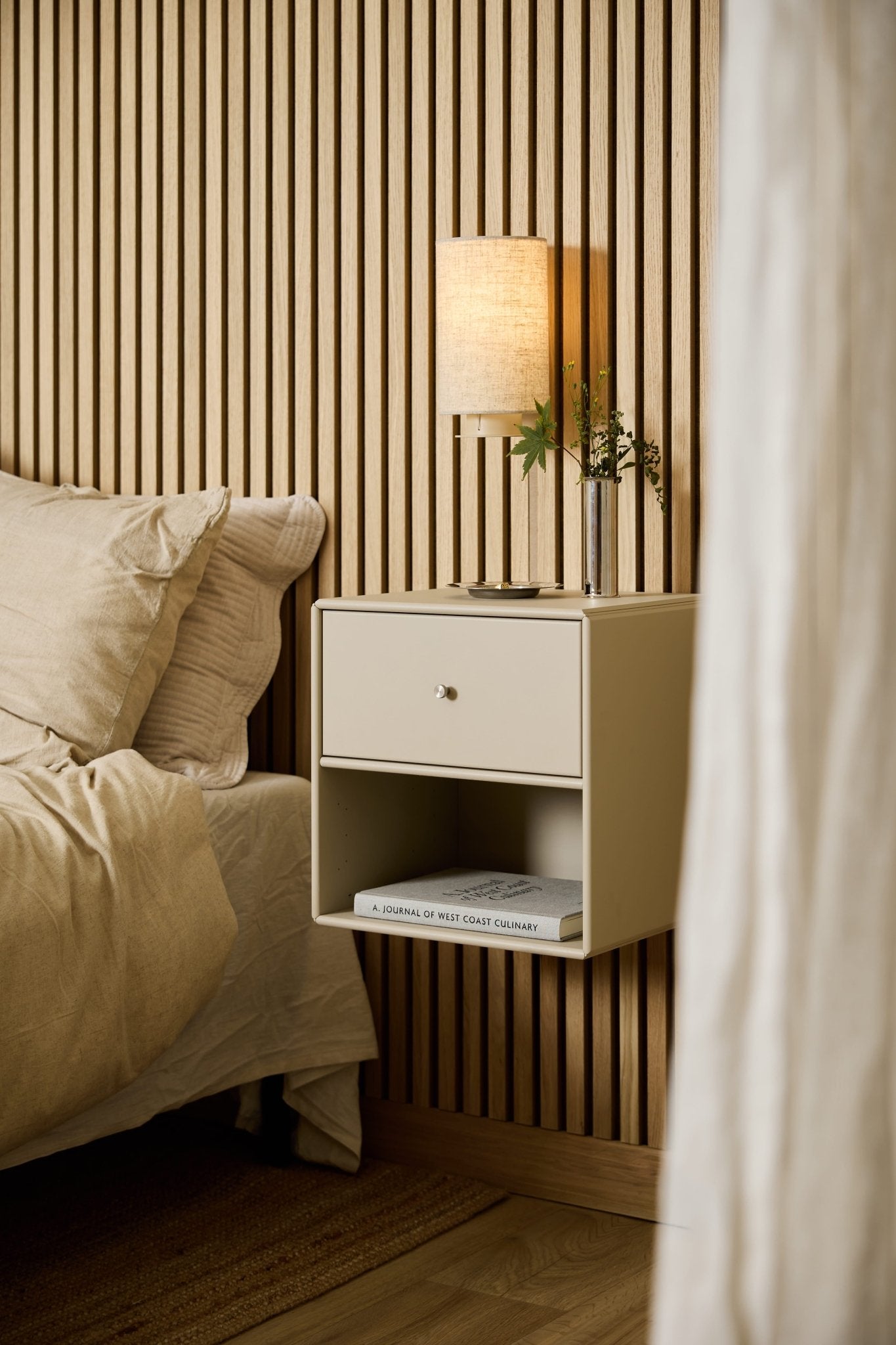 En beige WoodUpp Arka-bokhylla med låda vid sängen rymmer en lampa, böcker och en vas och står framför en WoodUpp Akupanel – en akustisk väggpanel av träribbor.