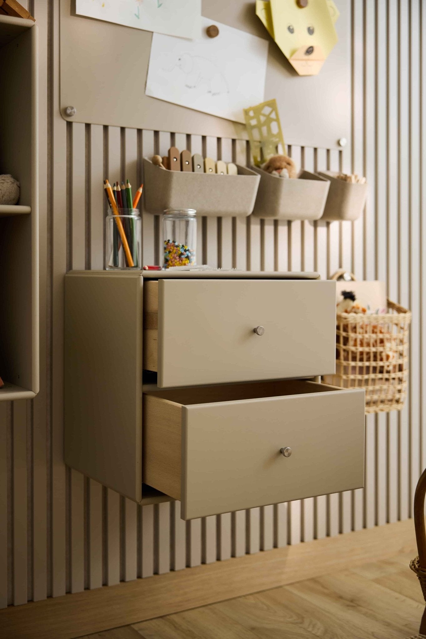 Två beige Arka-bokhyllor med lådor från WoodUpp, delvis öppna, där konstmaterial och leksaker förvaras; i närheten finns slutna förvaringsutrymmen och korgar.