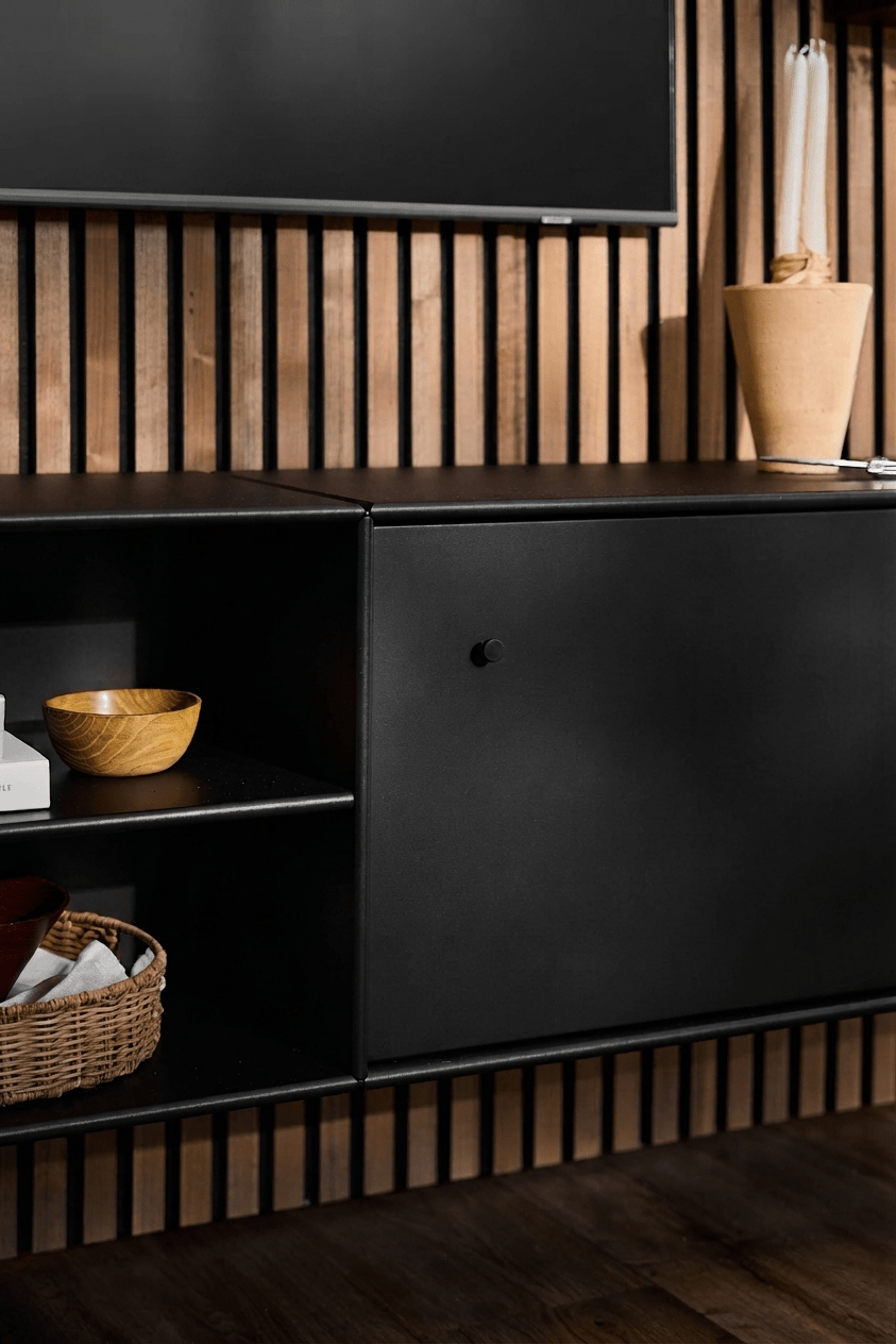 Svart TV-bänk från WoodUpp Arka med öppna hyllor för böcker och korgar, en beige vas, smart kabelhantering och en vertikal Akupanel-akustikpanel i trä som bakgrund.