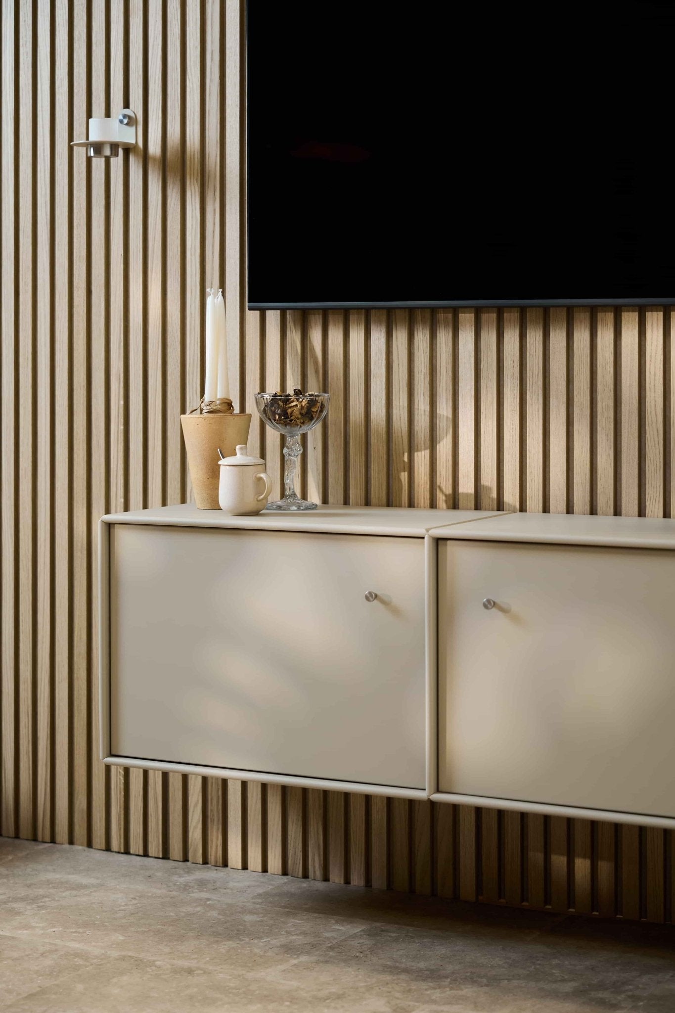Ett minimalistiskt vardagsrum med TV-bänken WoodUpp Arka i beige, en vägg med akustiska träpaneler från Akupanel, en väggmonterad TV, ljus, en glasskål, en vit burk och snyggt ordnade kablar.