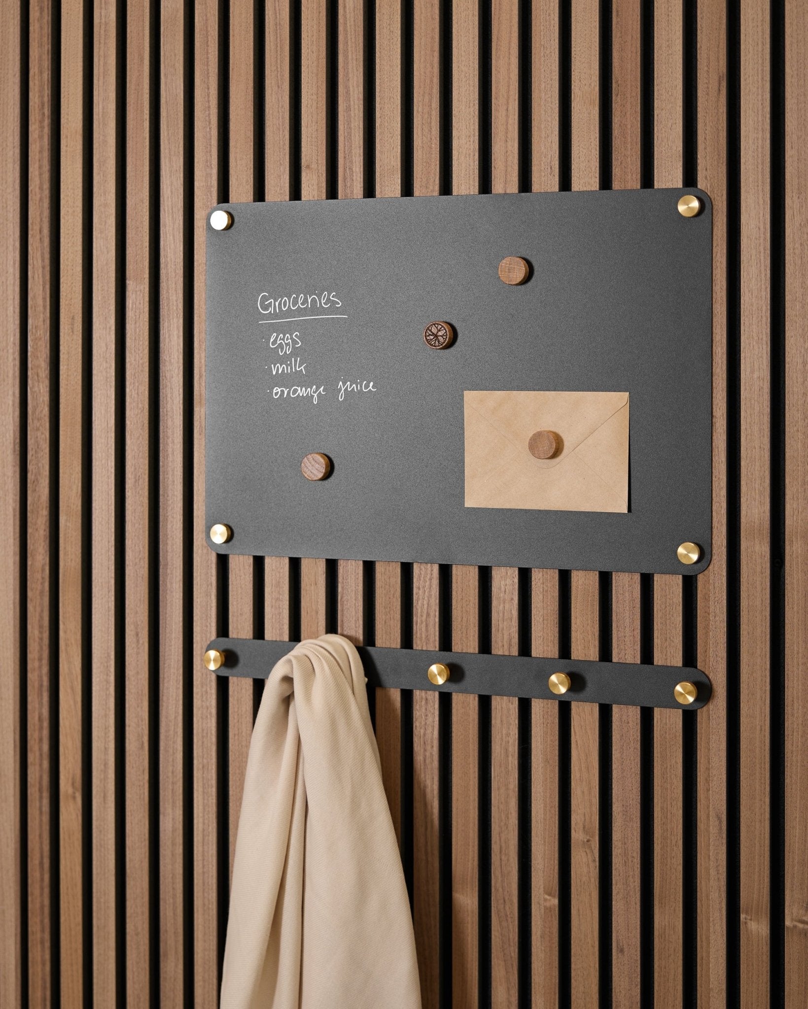 En small black Norr magnetisk tavla från WoodUpp rymmer en inköpslista, ett kuvert, magneter och beige tyg på krokar, monterad på en Akupanel Akustikpanel.