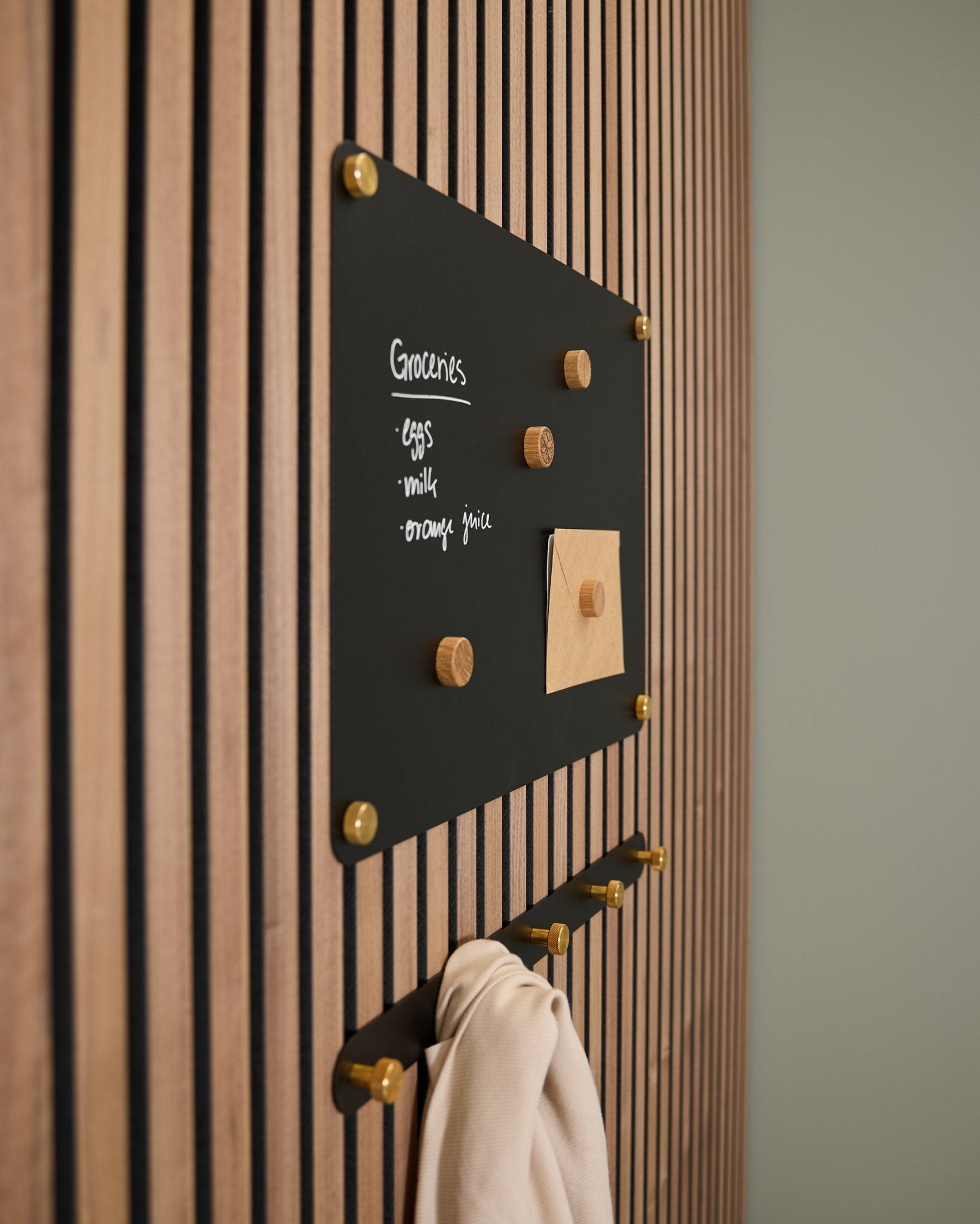 En small black Norr magnetisk tavla från WoodUpp, monterad på en Akupanel akustisk träpanel, visar en inköpslista och en anteckning, med krokar nedtill som håller ett ljust tyg.