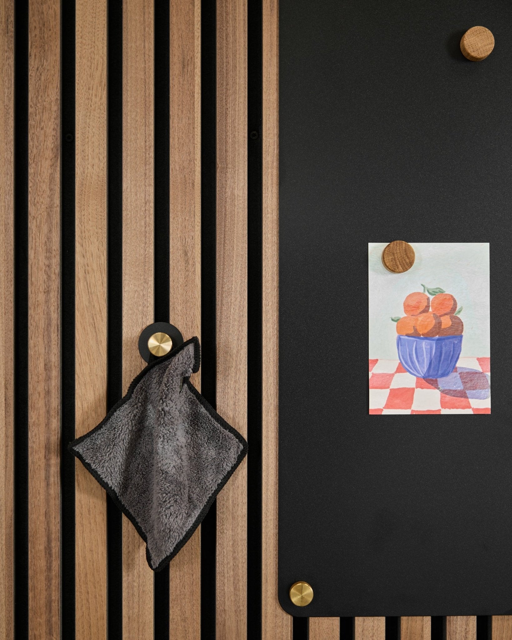 En small handduk hänger på en nordisk akustisk ribbpanel bredvid en black tavla som visar en teckning av apelsiner i en skål, fäst med två guldfärgade magneter.