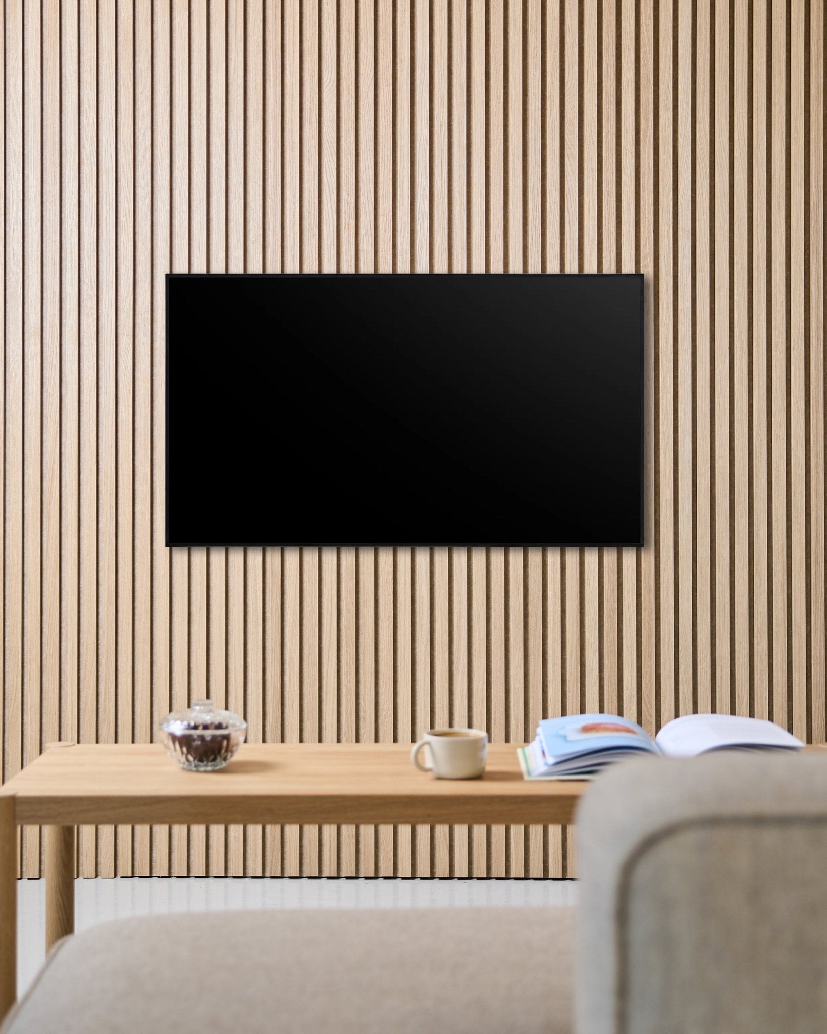 Ett Fjell TV-fäste (small) från WoodUpp håller en platt-tv på en nordisk Akupanel akustikvägg med trälameller, ovanför ett bord med kopp, skål och ett uppslaget magasin bredvid en beige soffa.
