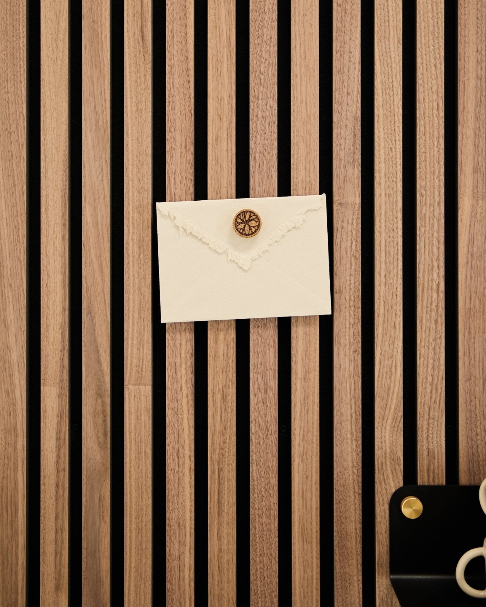 Ett krämfärgat kuvert med lacksigill är fäst på en akustisk slatwallpanel i ek från WoodUpp med Norr Poster Magnet set i Natural Oak; sax och bricka finns i närheten.