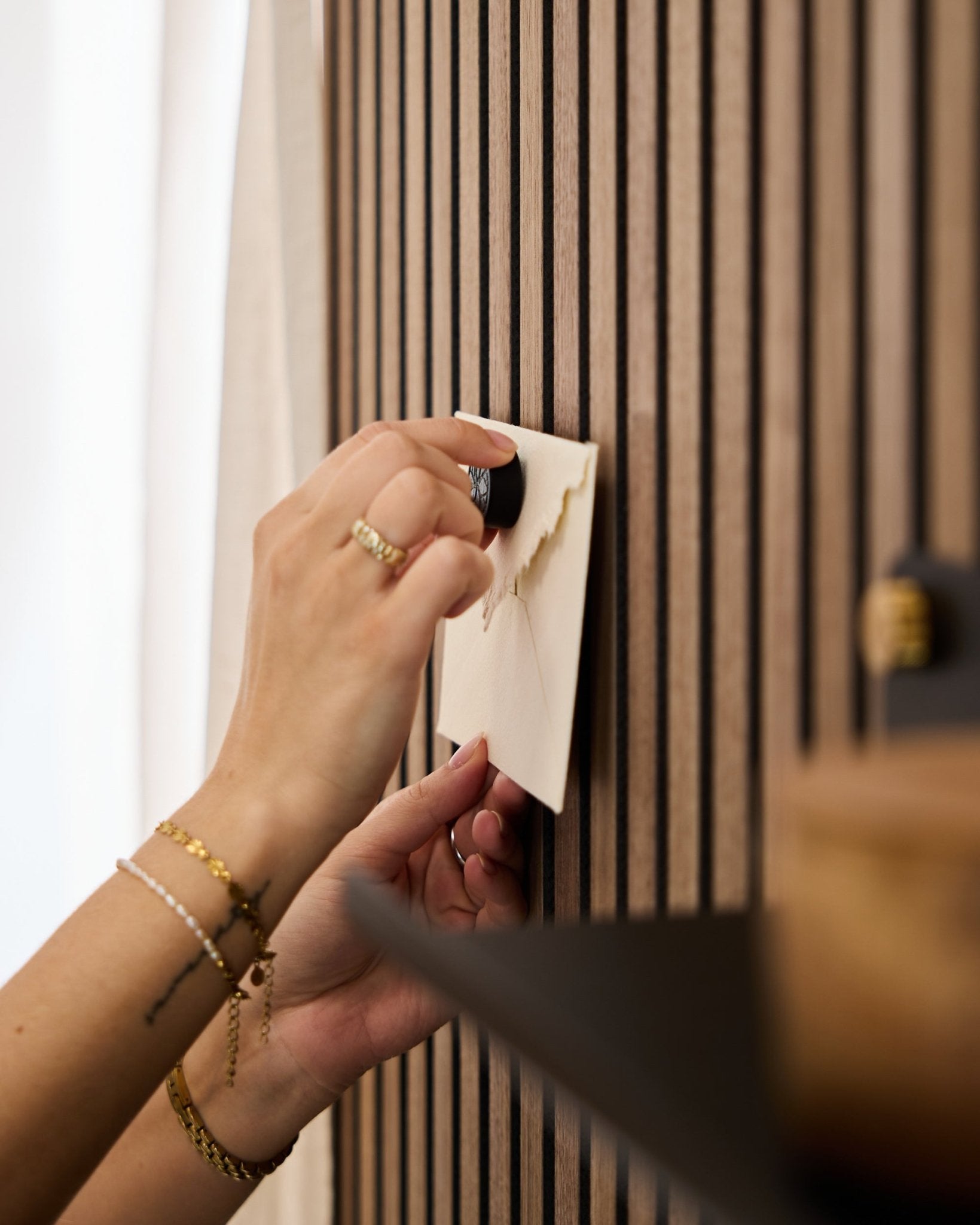 En person med guldfärgade armband och ringar fäster ett kuvert på en akustisk slatwall-panel i ek från WoodUpp med Norr Poster Magnet-setet i Black Oak.