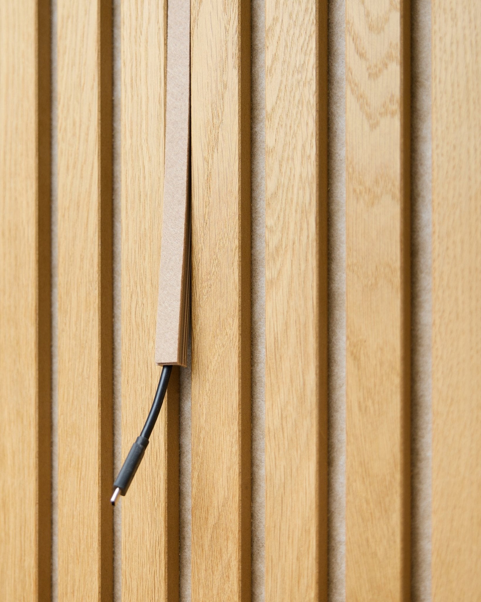 En black kabel med kabelhantering är delvis dold i en WoodUpp Dupps Beige Pop-on Cable Cover, monterad på vertical Akupanel akustiska trälameller.