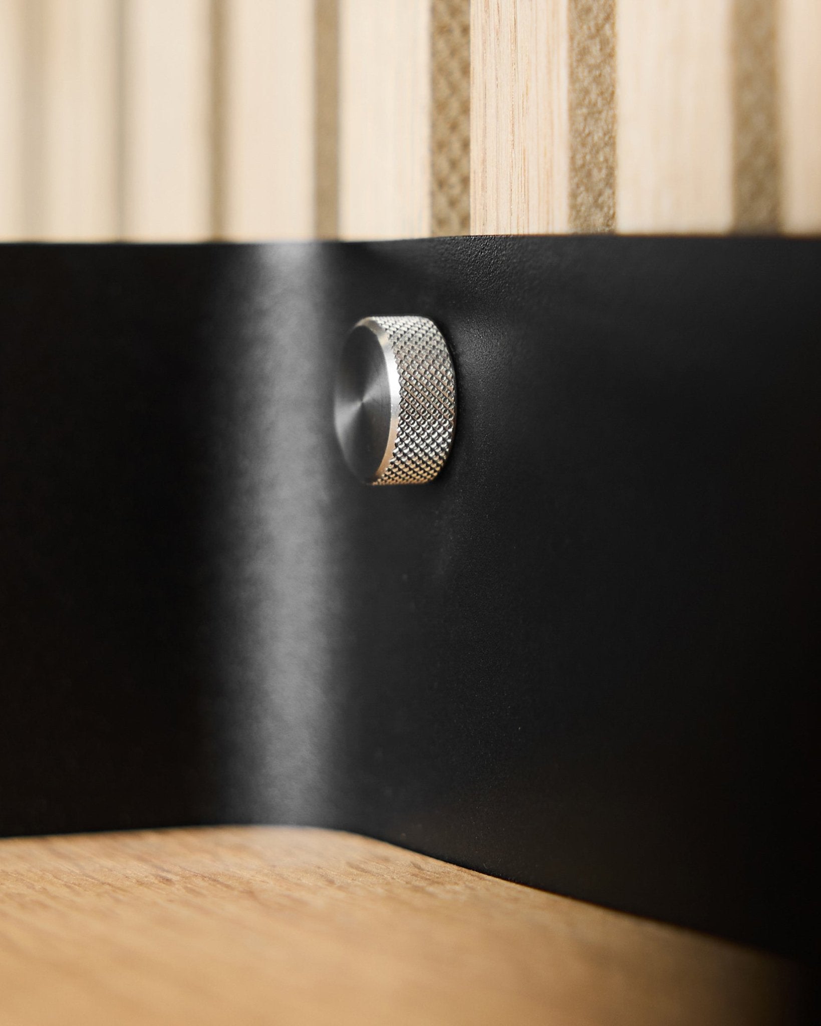 Närbild på två WoodUpp fingerskruvar (M5x10 mm, 20x8.8 i rostfritt steel) på en black metallyta med Akupanel akustiska trälameller i bakgrunden.
