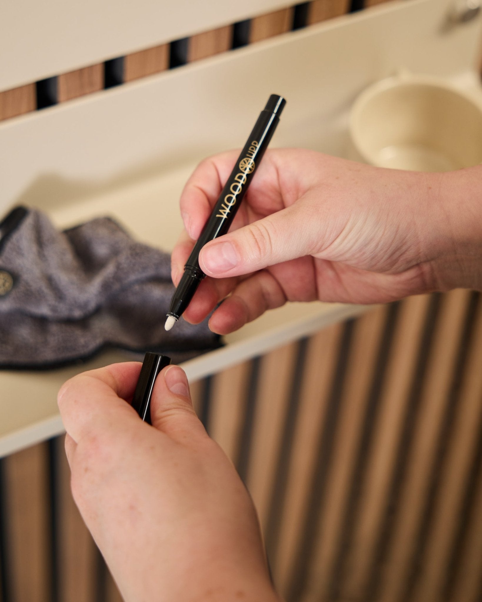 En person håller i en öppen WoodUpp Norr | Blackboard set-penna bredvid en mikrofiberduk och en beige kopp, produkter utformade för användning med akustiska träpaneler.