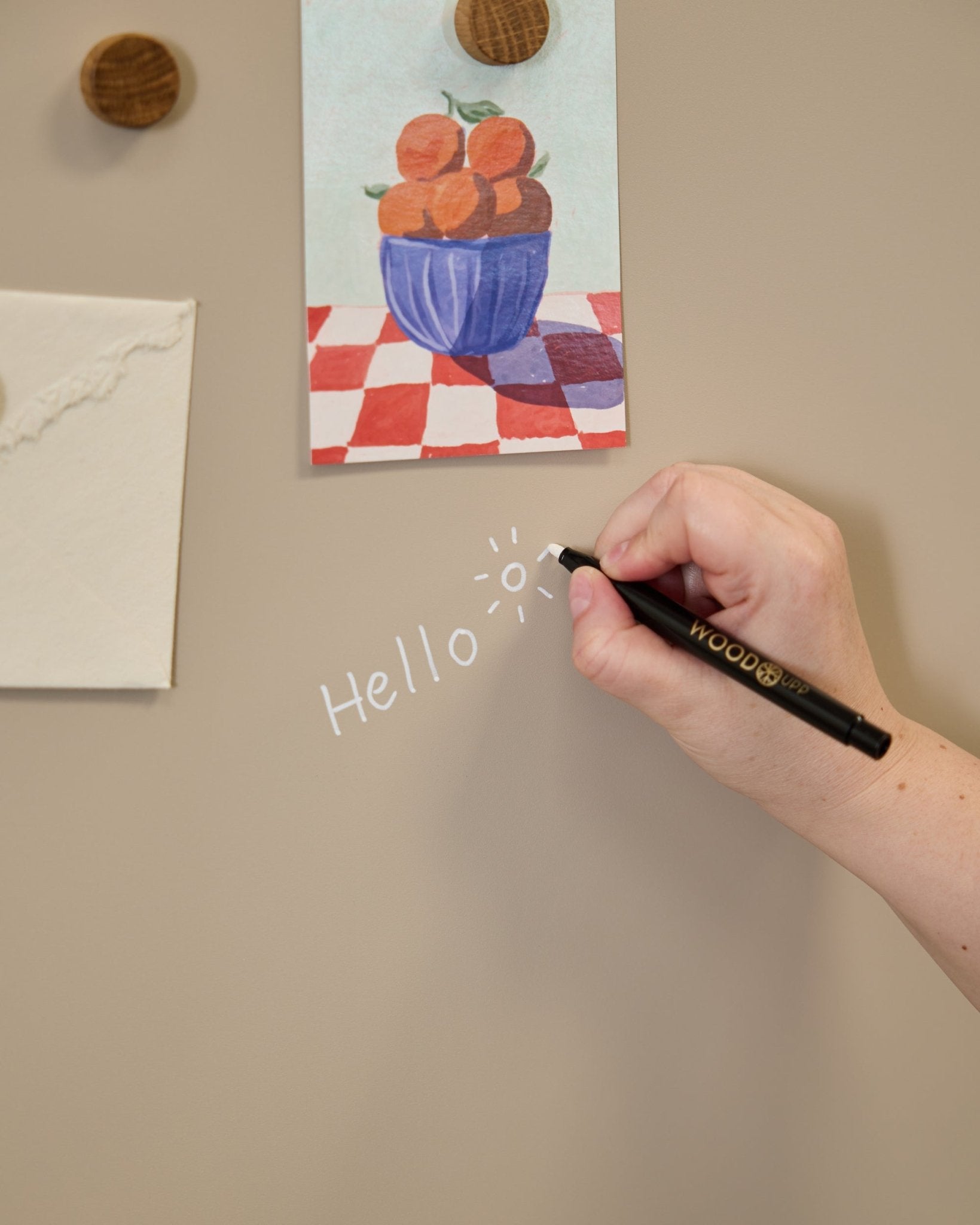 En hand skriver hello med en soldoodle på WoodUpps Norr Blackboard, placerad bredvid konst som fästs med runda magneter på en Akustikpanel.