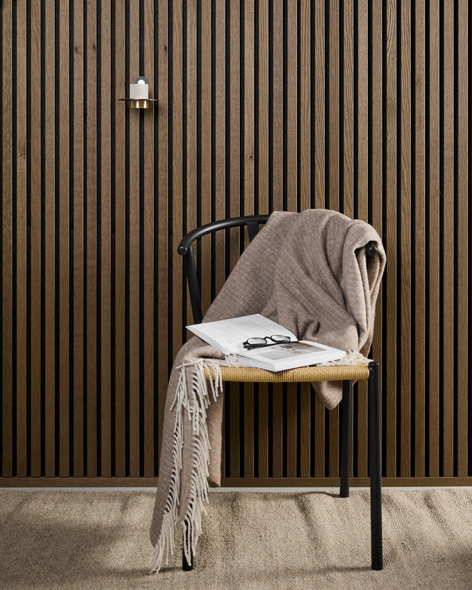 En modern stol med en beige pläd, en uppslagen bok och ett par glasögon står intill en Akupanel akustikpanel i trä samt en black och brass WoodUpp Norr ljusstake.