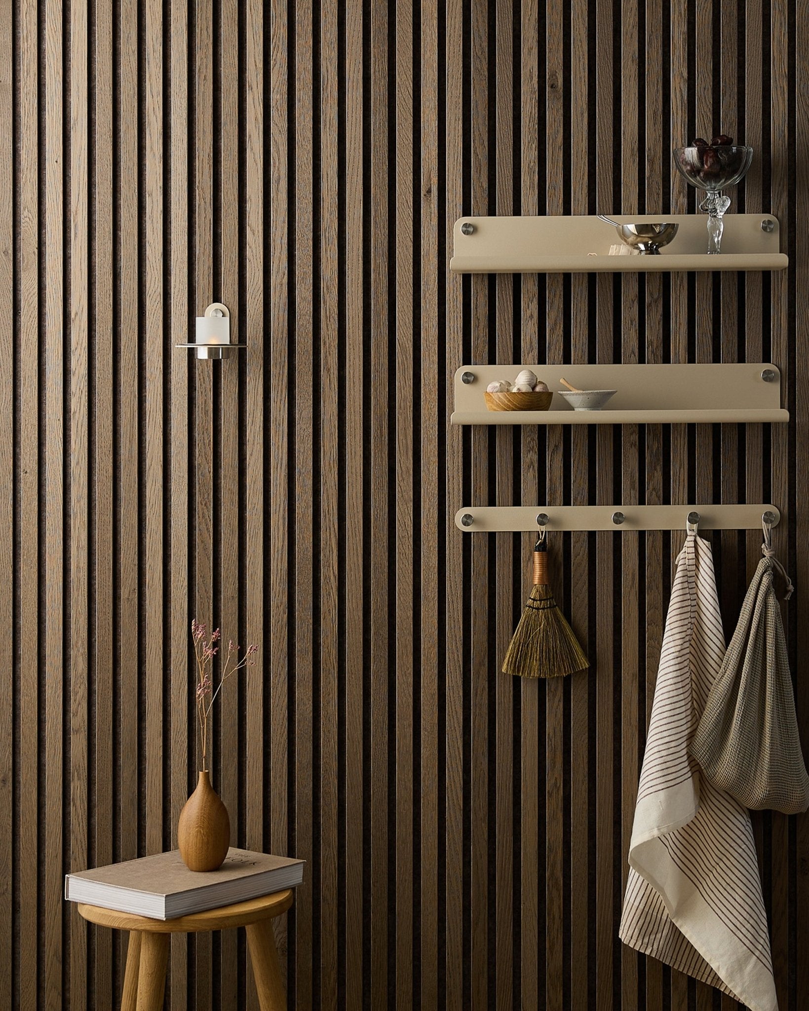 En träpall med torkade blommor står bredvid en Akupanel Akustikpanel från WoodUpp, intill beige hyllor med tallrikar, en borste, handdukar och en Norr Candle Holder.
