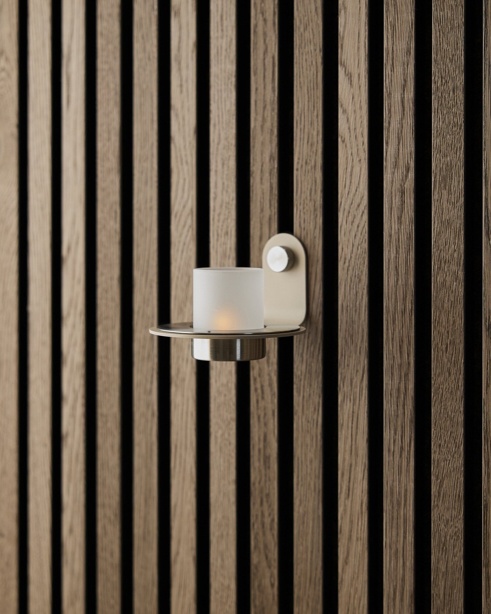 Ljusstaken Norr i beige rostfritt steel från WoodUpp, visad på en vägg med beige Akupanel Akustikpanel.