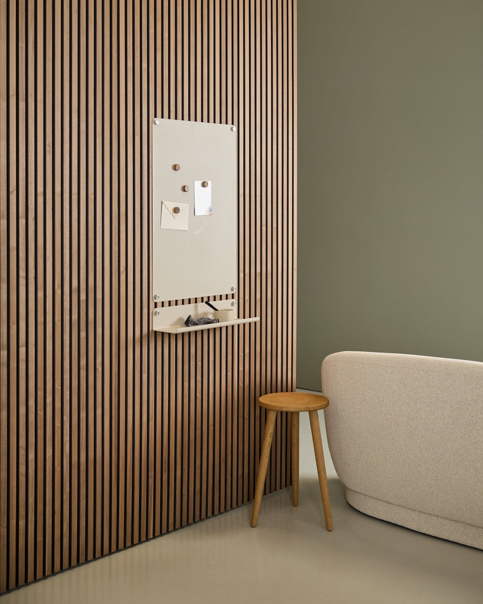 Minimalistisk interiör med beige soffa och träpall, med en large beige WoodUpp Norr magnettavla monterad på en nordisk Akupanel akustikpanel i trä intill en grön vägg.