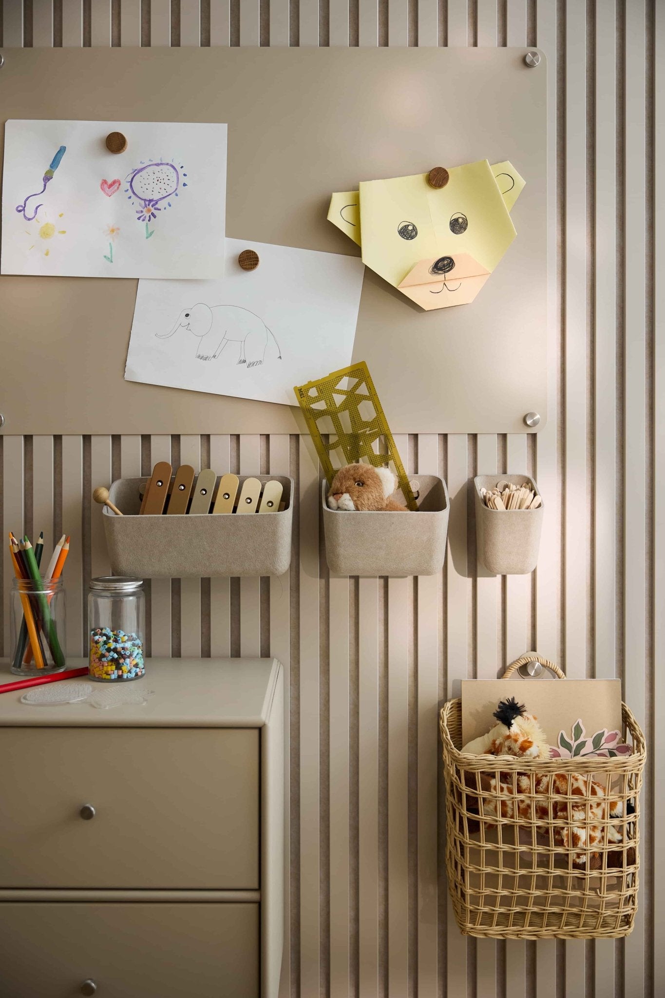 Barnens konstverk och leksaker är uppställda mot en beige vägg med en WoodUpp-akustikpanel, där det finns filtbehållare, korgar, en låda och en hålskiva.