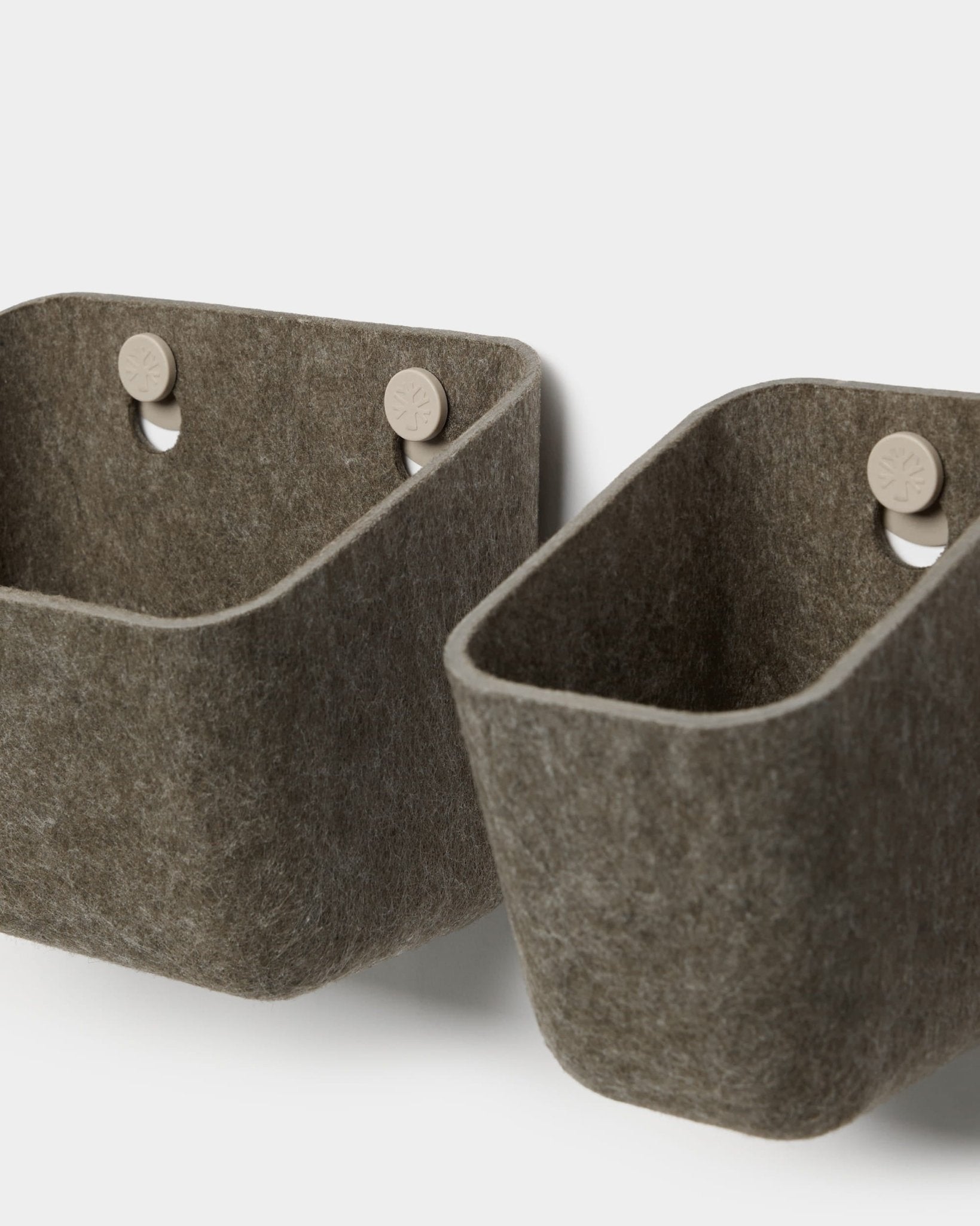 Två Luva-behållare i filt i mörk, varmgrå färg från WoodUpp hänger på beige krokar som är fästa vid en akustikpanel och bildar en minimalistisk väggförvaring med rundade hörn.