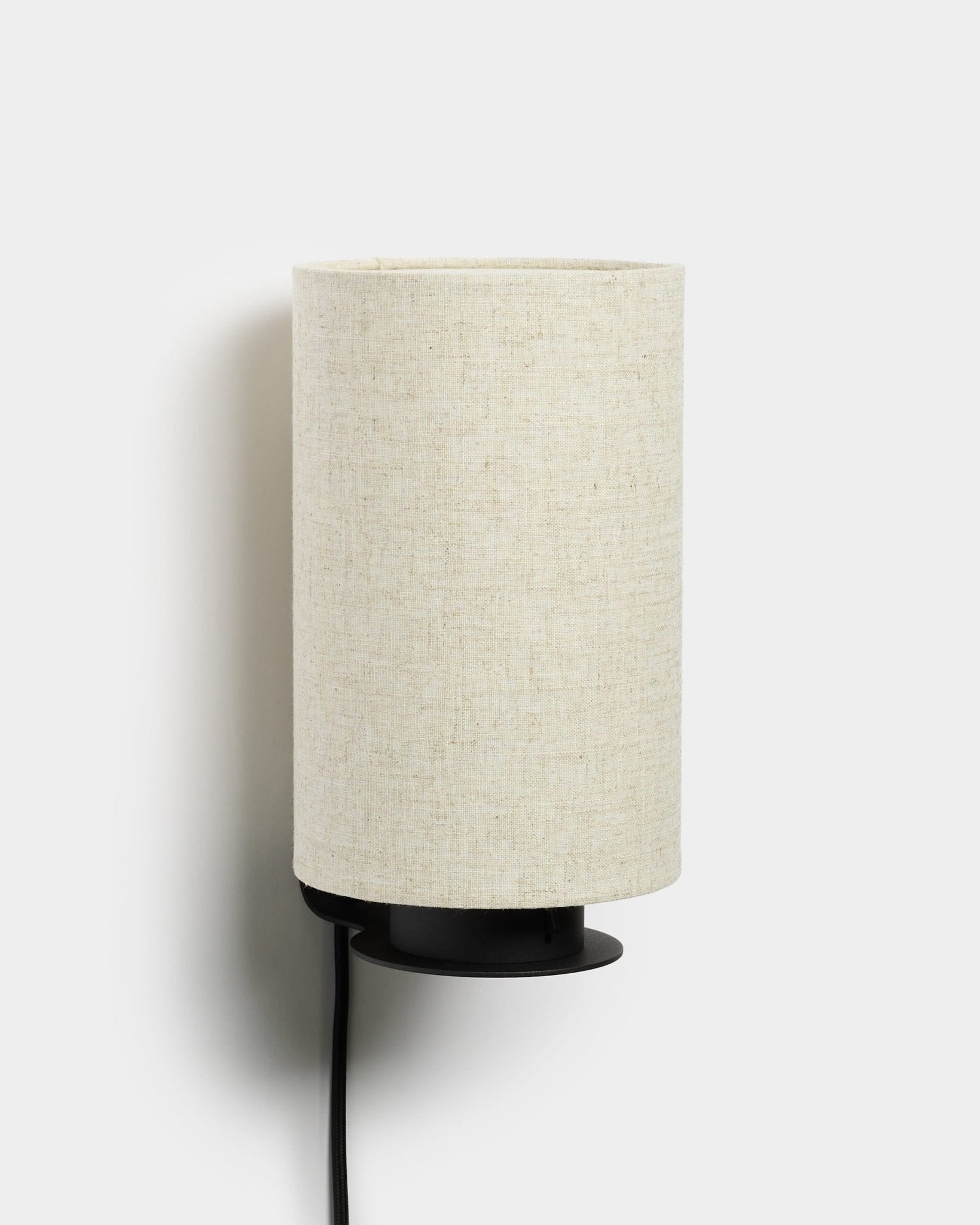 Feya vägglampa från WoodUpp med metallfot i black, texturerad skärm i linne-bomull i beige och synlig elkabel i black. Nordisk minimalistisk design.
